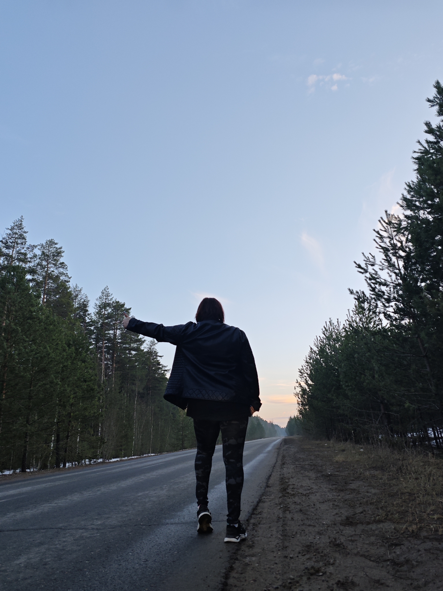 The image depicts a person standing on the side of a road in Chaikovsky, Russia. The person is facing away from the camera, with their left arm extended in what appears to be a gesture of hitchhiking. They are wearing a black jacket and patterned leggings, with dark shoes. The road is flanked by dense forest on both sides. The sky is a gradient of light blue with hints of pink and orange near the horizon, indicating either sunrise or sunset. The road surface appears wet, and there is some mud on the shoulder. The scene has an isolated, rural feel.