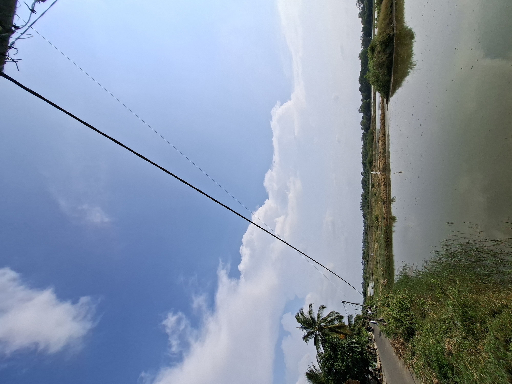 The image depicts a rural landscape under a bright blue sky with scattered white clouds. A power line stretches diagonally across the upper portion of the frame. To the left, a paved road runs alongside lush green vegetation and several palm trees. Beyond the road, a body of water, possibly a paddy field, reflects the sky and surrounding greenery. In the distance, a flat, open area with more vegetation and bodies of water stretches towards the horizon, with a line of trees forming a distant treeline. The scene appears to be in Kruengmate, Indonesia, based on the provided context. There are no people or active interactions visible.