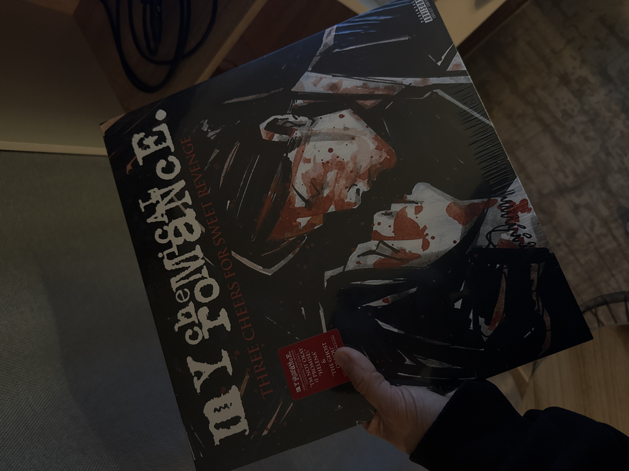A person is holding a vinyl record album titled "My Chemical Romance: Three Cheers for Sweet Revenge". The album cover features an illustration of two figures covered in red splatters, presumably blood. A sticker on the album highlights the songs "I'm Not Okay (I Promise)", "Helena", and "The Ghost of You". The background is out of focus, showing part of a wooden shelf with black cables. There is no indication that the image was taken in Ukromne, Ukraine.