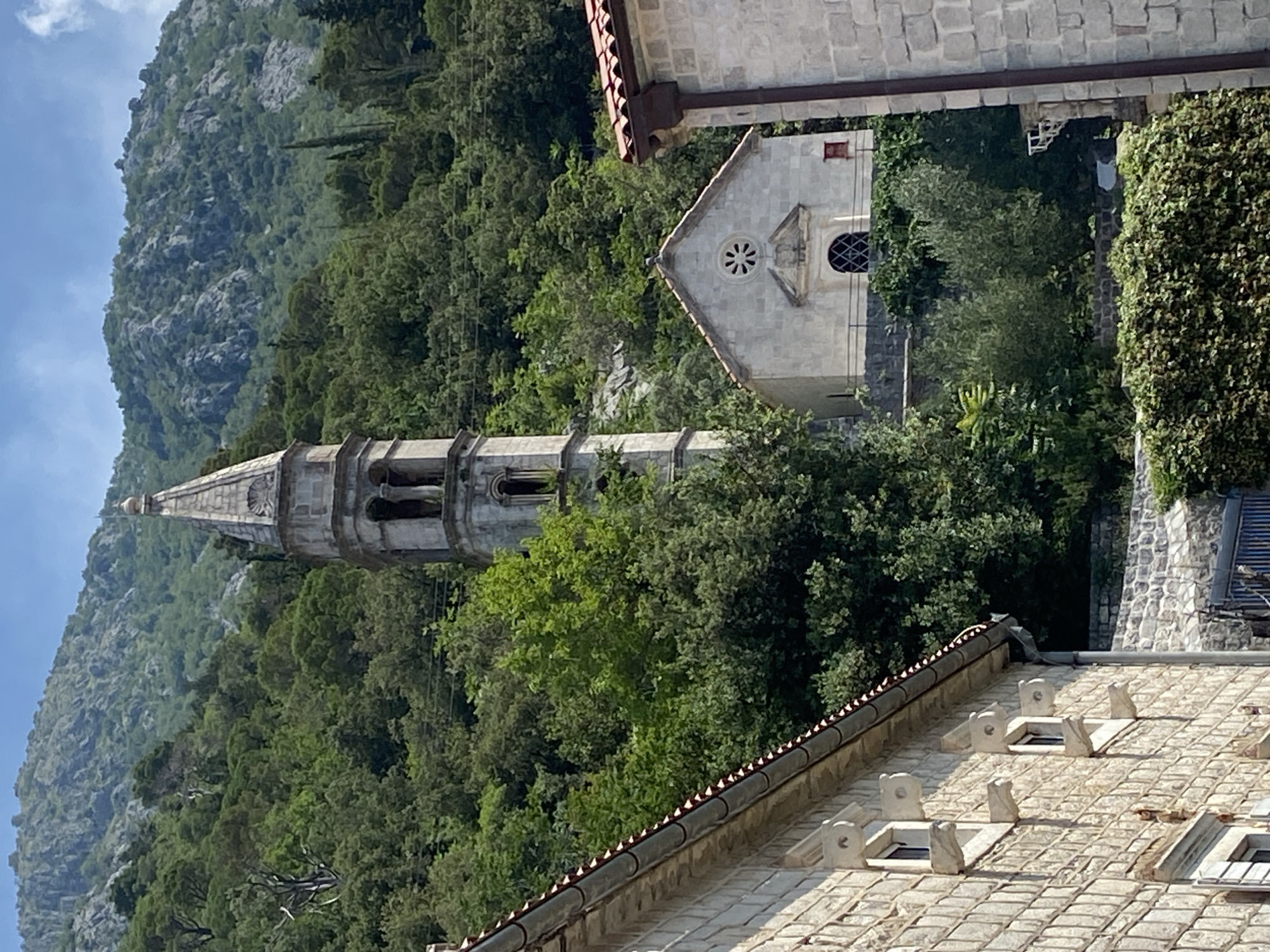 The image shows a bell tower and a small church with a stone facade, nestled on a hillside covered in lush green trees. The architecture suggests a historic European setting, likely in the Mediterranean region. In the foreground, a portion of a stone building with visible windows and a red-tiled roof is visible on the left side of the frame, and another stone building with a red-tiled roof is on the right. The bell tower is tall and slender, with arched openings and a pointed spire. The church is a single-story structure with a distinct rose window above an arched doorway. The background is dominated by a steep, rocky mountain, also covered in dense vegetation. The sky is clear and blue, suggesting a sunny day. The scene is quiet and serene, evoking a sense of peace and timelessness, characteristic of old coastal towns. Based on the style of architecture and the mountainous coastal landscape, this scene is likely located in Perast, Montenegro, a town known for its Baroque architecture and prominent bell towers overlooking the Bay of Kotor.