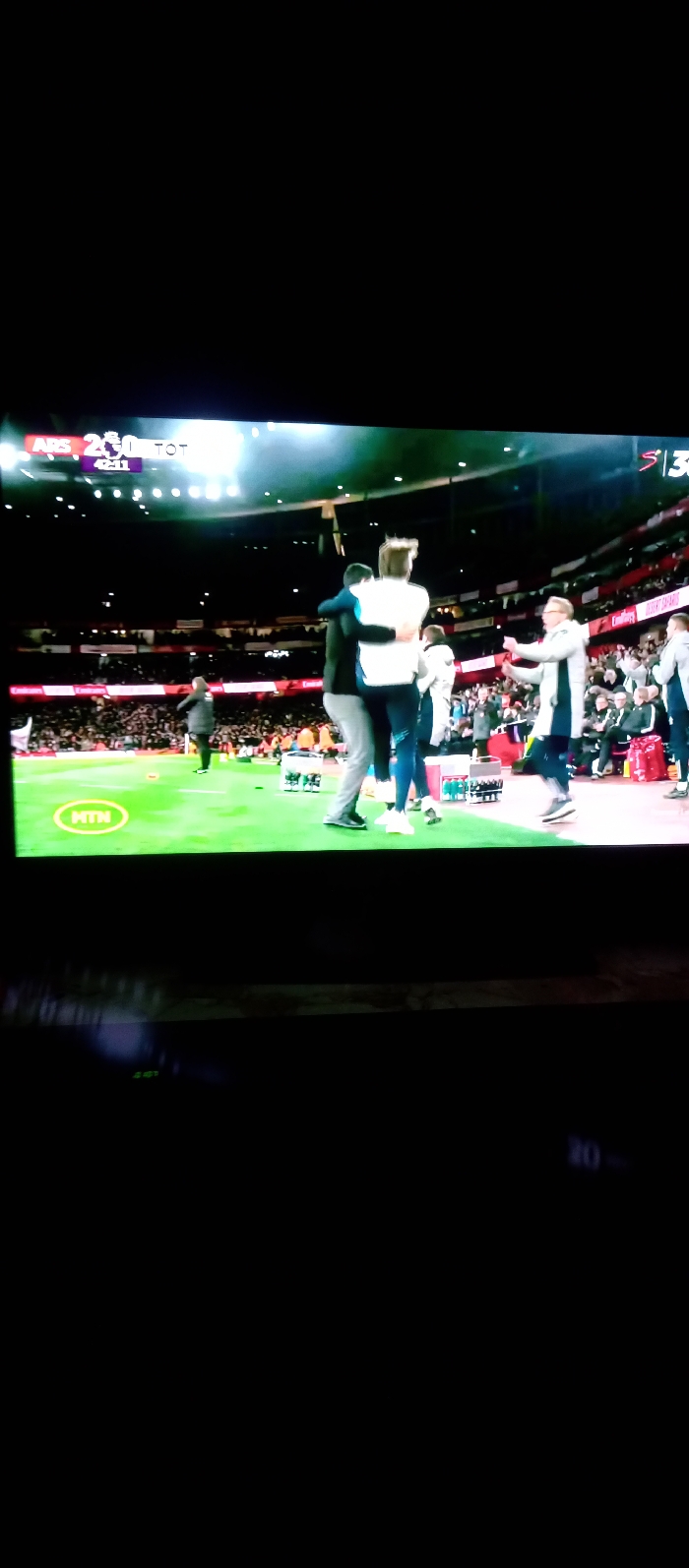 A TV screen displays a live soccer match between Arsenal (ARS) and Tottenham (TOT), with the score 2-0 at 42:11. On the sidelines, team members are celebrating, embracing each other. The stadium lights shine brightly, illuminating the field and the stands filled with spectators. A green MTN logo is visible on the field.