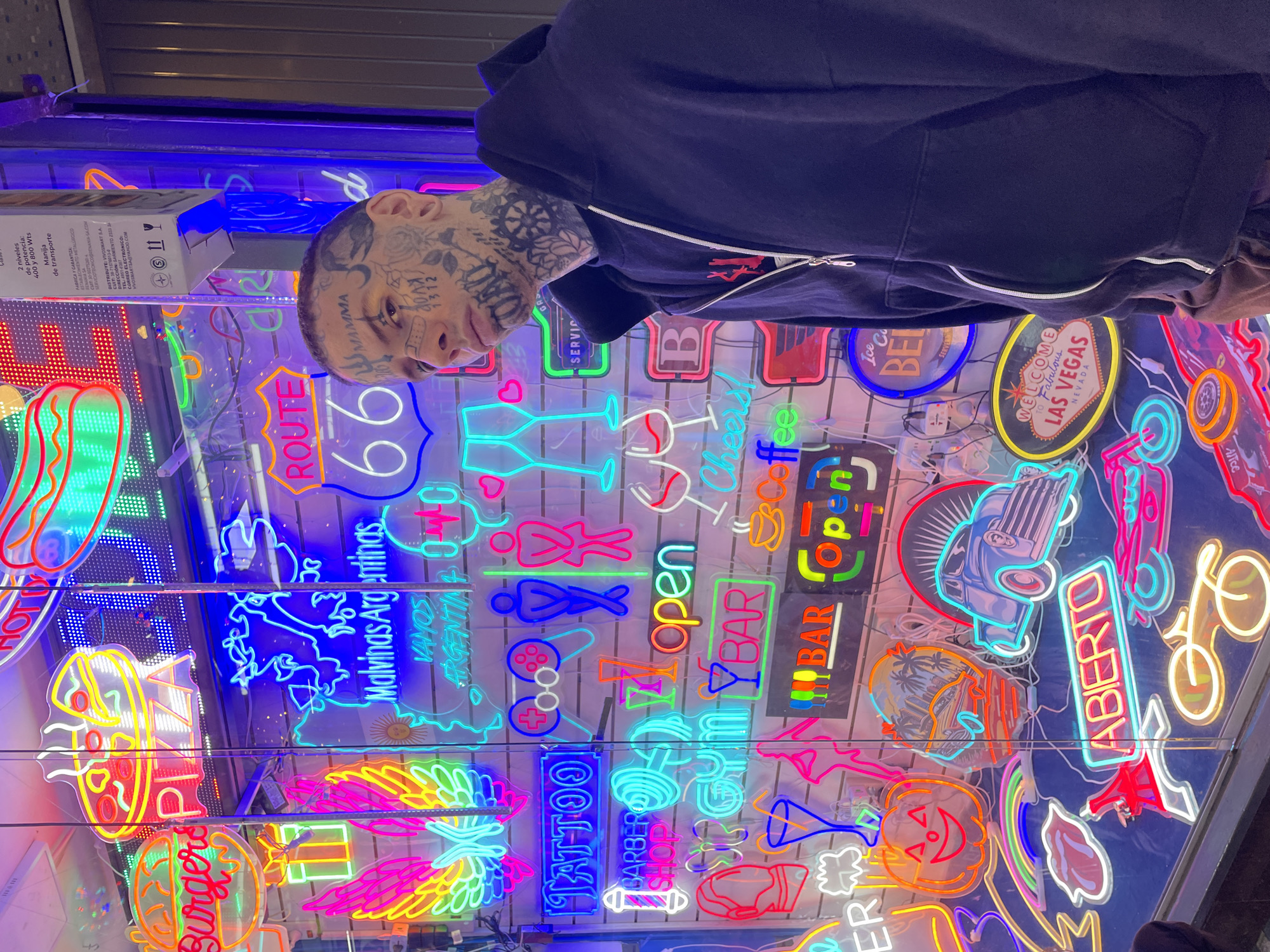A person with extensive facial and head tattoos, wearing a dark blue hooded sweatshirt, is captured in a close-up shot, looking directly at the camera. The image is rotated ninety degrees clockwise. The person's face shows tattoos including "MAMMA" and "KVD".
The background is a vibrant, dense display of numerous glowing neon signs, suggesting an indoor shop or market specializing in such items. The neon signs feature a wide array of words, symbols, and shapes in various bright colors like red, blue, green, yellow, and pink.
Visible text on the neon signs includes "HOTT," "Burgers," "ROUTE 66," "Malvinas Argentinas," "VAMOS ARGENTINA," "TATTOO," "BARBER SHOP," "open," "BAR," "Cheers," "ABIERTO" (Spanish for "Open"), and "WELCOME TO FABULOUS LAS VEGAS NEVADA." Other signs depict items like cocktail glasses, coffee cups, cars, bicycles, and abstract designs.
A cardboard box is partially visible on the left side of the frame, displaying text in Spanish. This text includes specifications like "2 niveles de potencia: 400 y 800 Wts," "Manija de transporte" (handle for transport), and company details for "VIVOSMART SA," including an address on "SARMIENTO 2333" and an email. The overall scene is brightly lit by the neon lights, creating a lively and colorful atmosphere in what appears to be a commercial setting in Buenos Aires, Argentina.