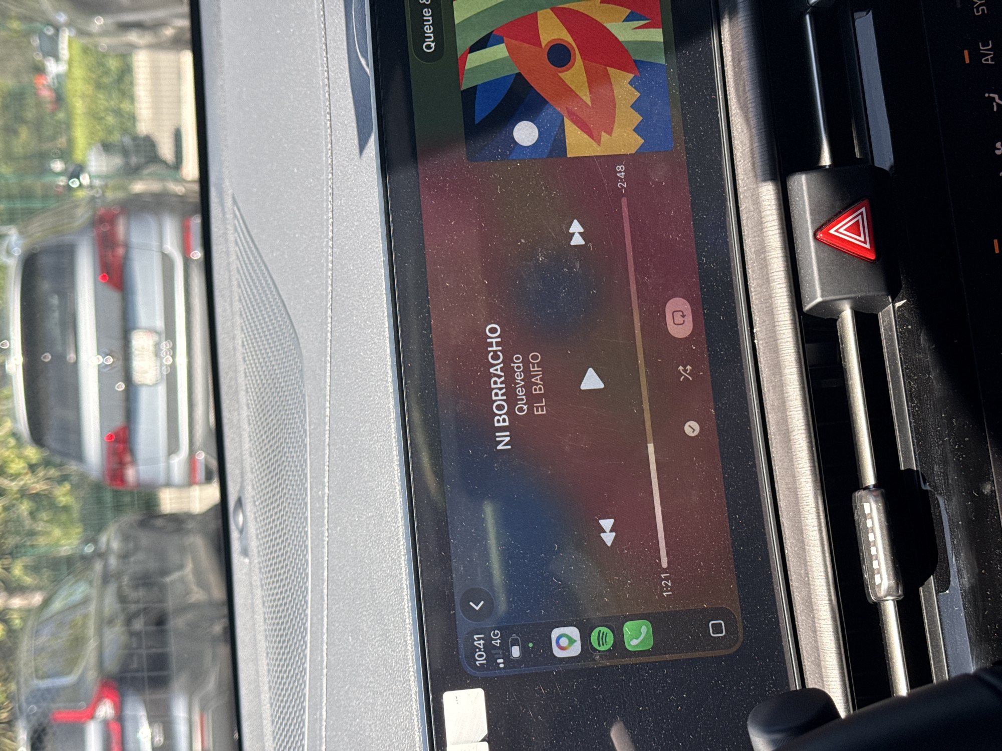 The image shows the infotainment screen of a car, displaying the Spotify app playing the song "Ni Borracho" by Quevedo and El Baifo. The time is 10:41 and the phone is connected via 4G. To the right of the screen are the car's hazard lights button and air conditioning vents. Outside the car window, there are blurry images of other vehicles and greenery, suggesting a street scene. The location cannot be determined to be in the city of Puebla, Mexico, without additional information.
