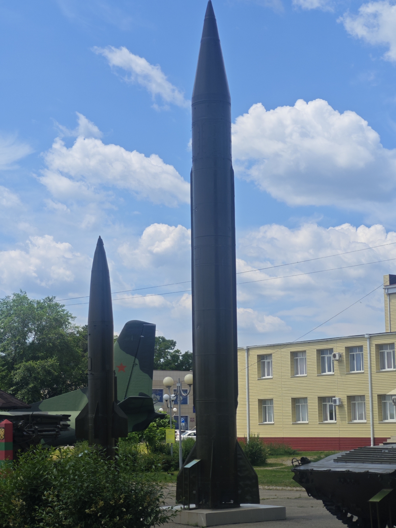 This is a full-shot, eye-level, outdoor photograph of a military display under a partly cloudy sky in the city of Vyselki, Russia. The main subjects are two large missiles, one standing upright in the foreground and another partially obscured to its left. The upright missile is dark grey and tapers to a sharp point at the top. The missile to the left has a camouflage pattern of green and black and a red star visible on its tail fin. Behind the missiles, there is a yellow building with multiple windows and air conditioning units. To the far right of the frame, a portion of a military vehicle, possibly a tank, is visible. Lush green bushes and trees are present in the foreground and midground, with power lines and a streetlamp also visible. The overall scene suggests a memorial or museum exhibit of military hardware.
