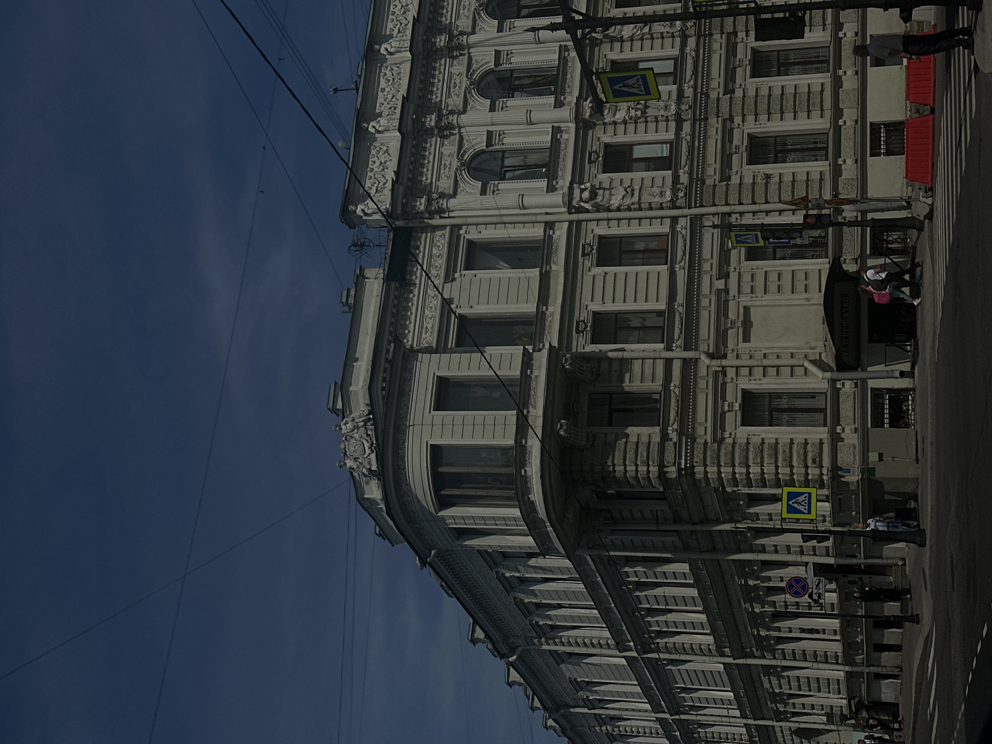 An external, medium-angle, ground-level shot captures a corner of a grand, ornate, multi-story building in a city. The building's facade is detailed with intricate architectural elements, including decorative carvings, columns, and arched windows, painted a pale, weathered grey. The sky above is a deep, dark blue, with visible overhead wires crisscrossing the frame. On the sidewalk in front of the building, pedestrians are visible. A crosswalk with white markings is present in the foreground. Traffic signals and street signs are also installed along the street. Two red plastic barriers are placed on the sidewalk. The lighting suggests it is daytime, with shadows indicating the sun's position.