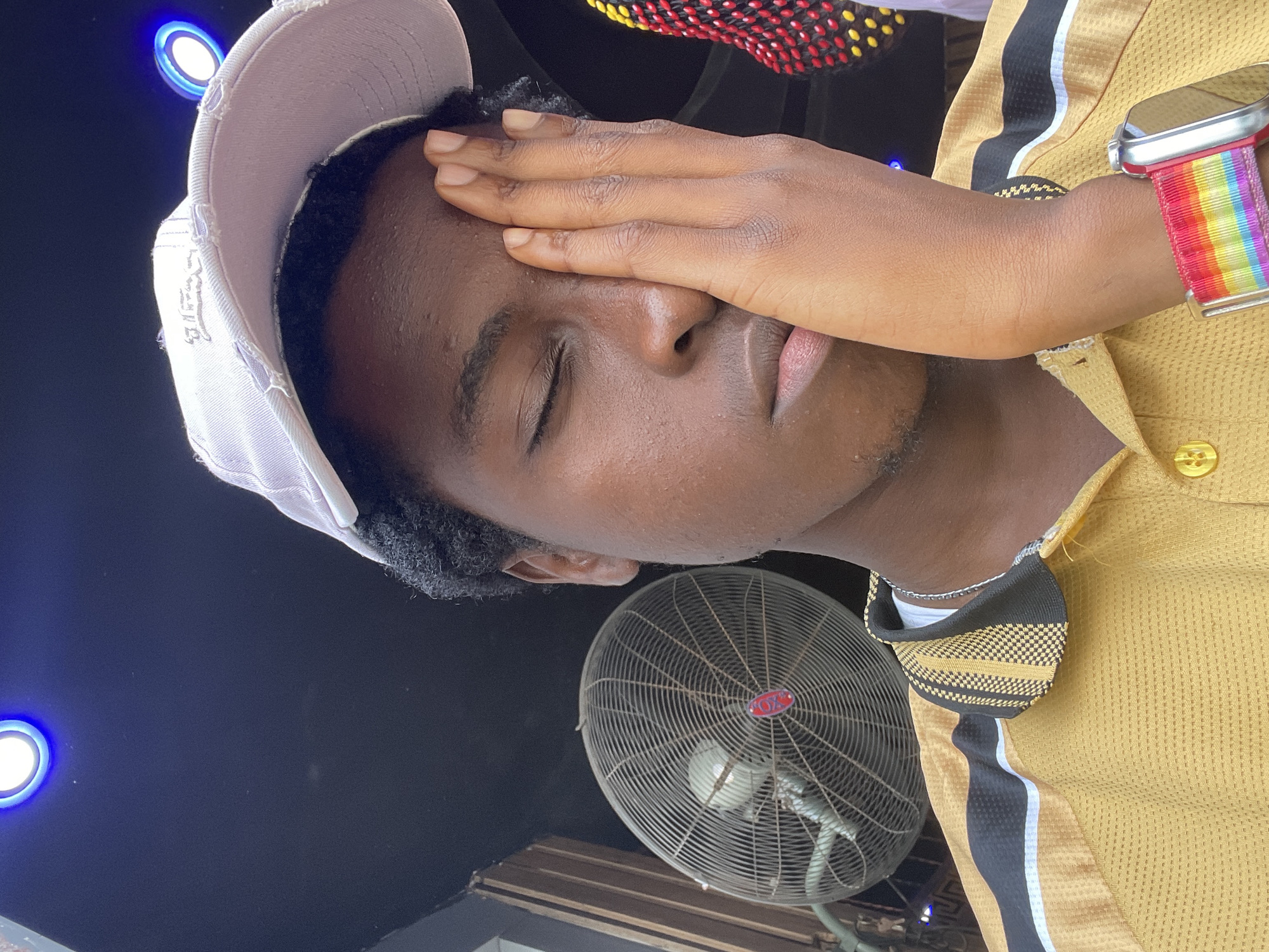 The image shows a close-up, sideways shot of a young Black person. They are wearing a light purple baseball cap and a yellow polo shirt. Their right hand is covering their eye, with their fingers resting on their forehead. The person has their eyes closed, suggesting they might be tired or shielding themselves from bright light. On their wrist, they are wearing a smartwatch with a rainbow-colored strap.
In the background, a large, metal industrial fan is visible. To the left of the frame, there are two blue LED lights. The overall setting appears to be indoors, possibly a shop or a workshop, given the presence of the fan and the lighting. The lighting suggests it might be daytime or an indoor space with artificial lights.
There is no visible text in the image. The subject's expression, with their hand over their eye and eyes closed, conveys a sense of discomfort or weariness. The location context is Nkpor, Nigeria.