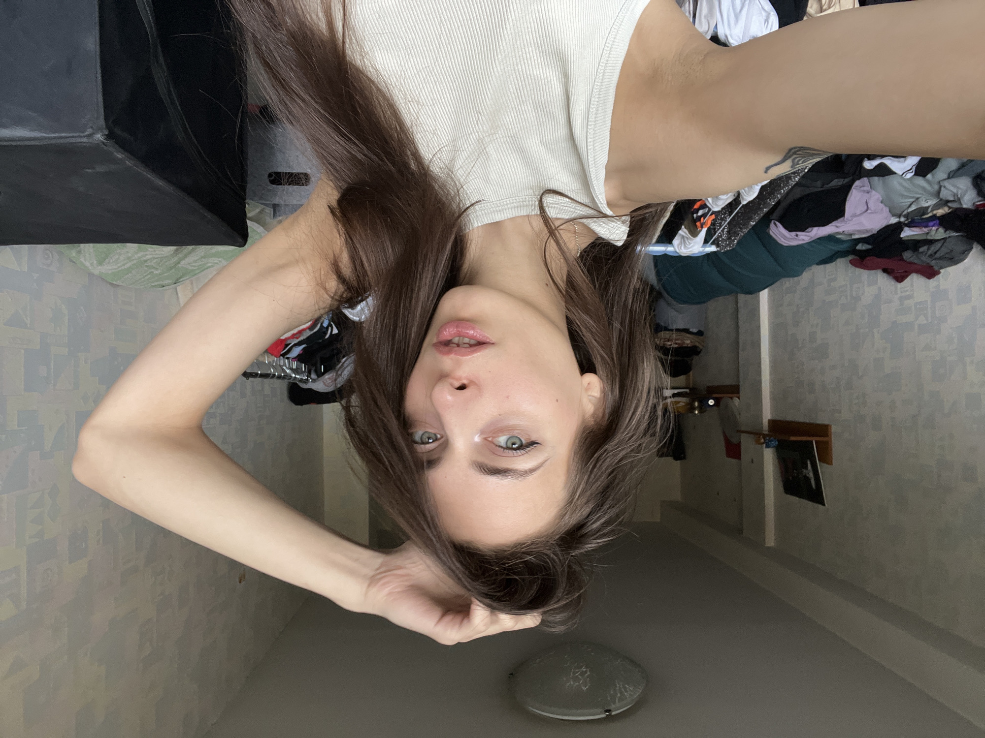 A young woman with long, dark hair and light eyes lies on her back, looking directly at the camera. She is wearing a white tank top. Her right arm is raised, with her hand touching her hair. The background is cluttered with clothes, a black box, and a patterned wall. A round, gray object is visible on the ceiling. The location appears to be an interior space, possibly a residential room. There is no information to suggest that the image was taken in the city of Krasnoyarsk, Russia.