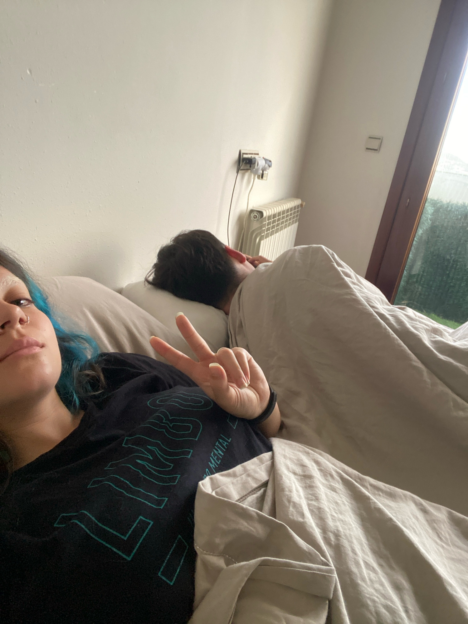 The image depicts a bedroom scene, likely in Gijón, Spain, with two individuals in bed. In the foreground, a person with blue-dyed hair is lying on their side, facing the camera. They are wearing a black t-shirt with teal lettering and have their left hand raised, displaying a peace sign with their index and middle fingers. Their right hand is resting on the blanket. Behind them, another person is sleeping, covered by a light-colored duvet. Their back is to the camera. The room features a white wall with a light switch and an electrical outlet visible. A radiator is mounted on the wall near a window, which appears to look out onto greenery. The lighting suggests it is daytime.