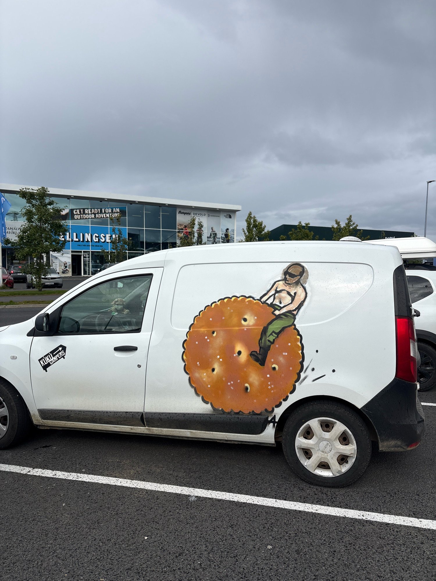A white van is parked in a parking lot in front of an outdoor equipment store in Reykjavik, Iceland. The van has a drawing of a man riding a giant cracker on its side. The store has large windows and signs advertising outdoor brands. The sky is cloudy and the ground is asphalt.
