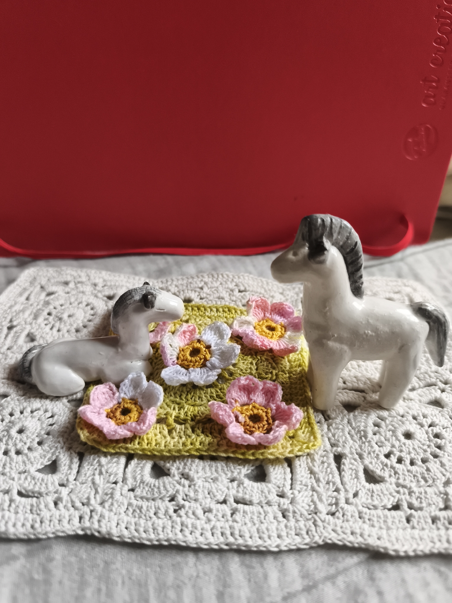 Two small ceramic horses, one lying down and one standing, are placed on a crocheted doily. In front of the horses, on a small square of yellow crochet, are three crocheted flowers, two pink and one white, each with a yellow center. In the background, a large, bright red object, possibly a book or a serving tray, is visible. The scene is lit with soft, natural light. The location in the city of Saint-Denis, France is not discernible from the image.