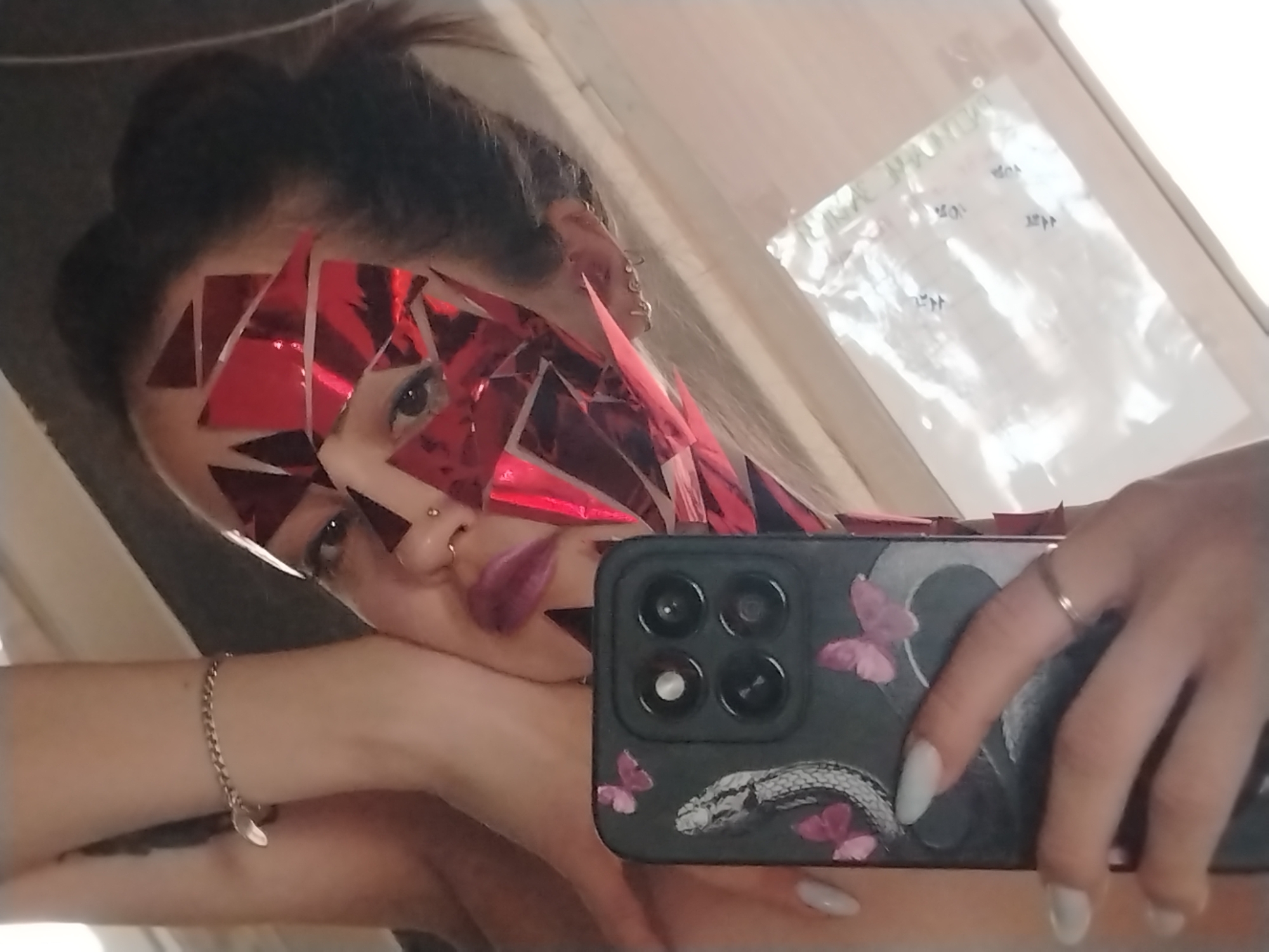 A woman is taking a selfie in a mirror. She has red geometric shapes glued to the right side of her face, covering her forehead, cheek, and jawline. She has a nose ring, multiple earrings, and her hair is pulled back in a bun. Her lips are painted a dark color. She is holding a smartphone with a black case decorated with a snake and butterflies. She is wearing a gold bracelet and a ring. The background is blurry and indistinct, showing a window and a light-colored wall. The location is Unknown, Unknown.