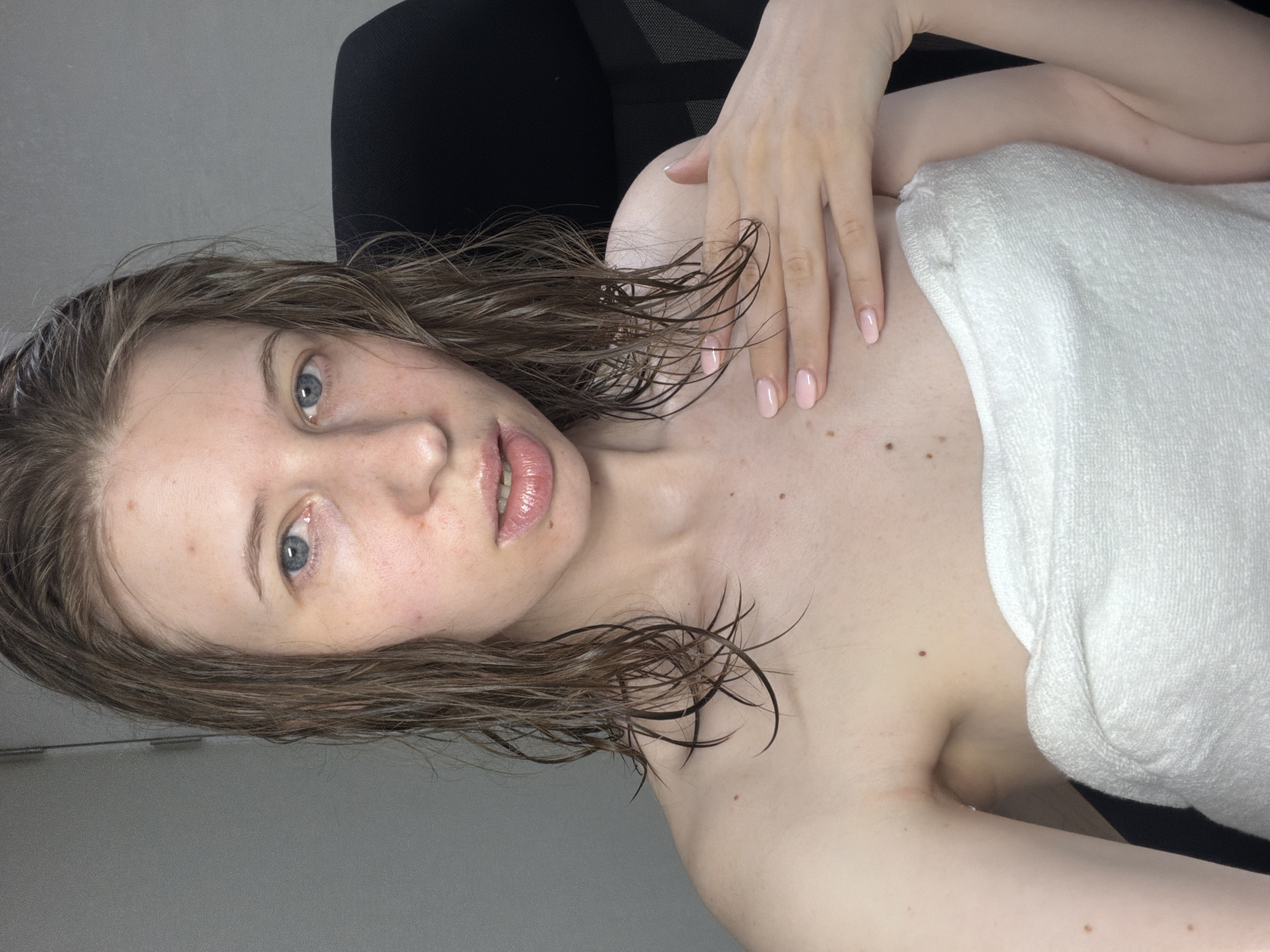 A young Caucasian woman with wet, brown hair is wrapped in a white towel and looking at the camera. She has blue eyes and is wearing light pink nail polish. The background is a plain gray wall. The woman appears to be indoors.