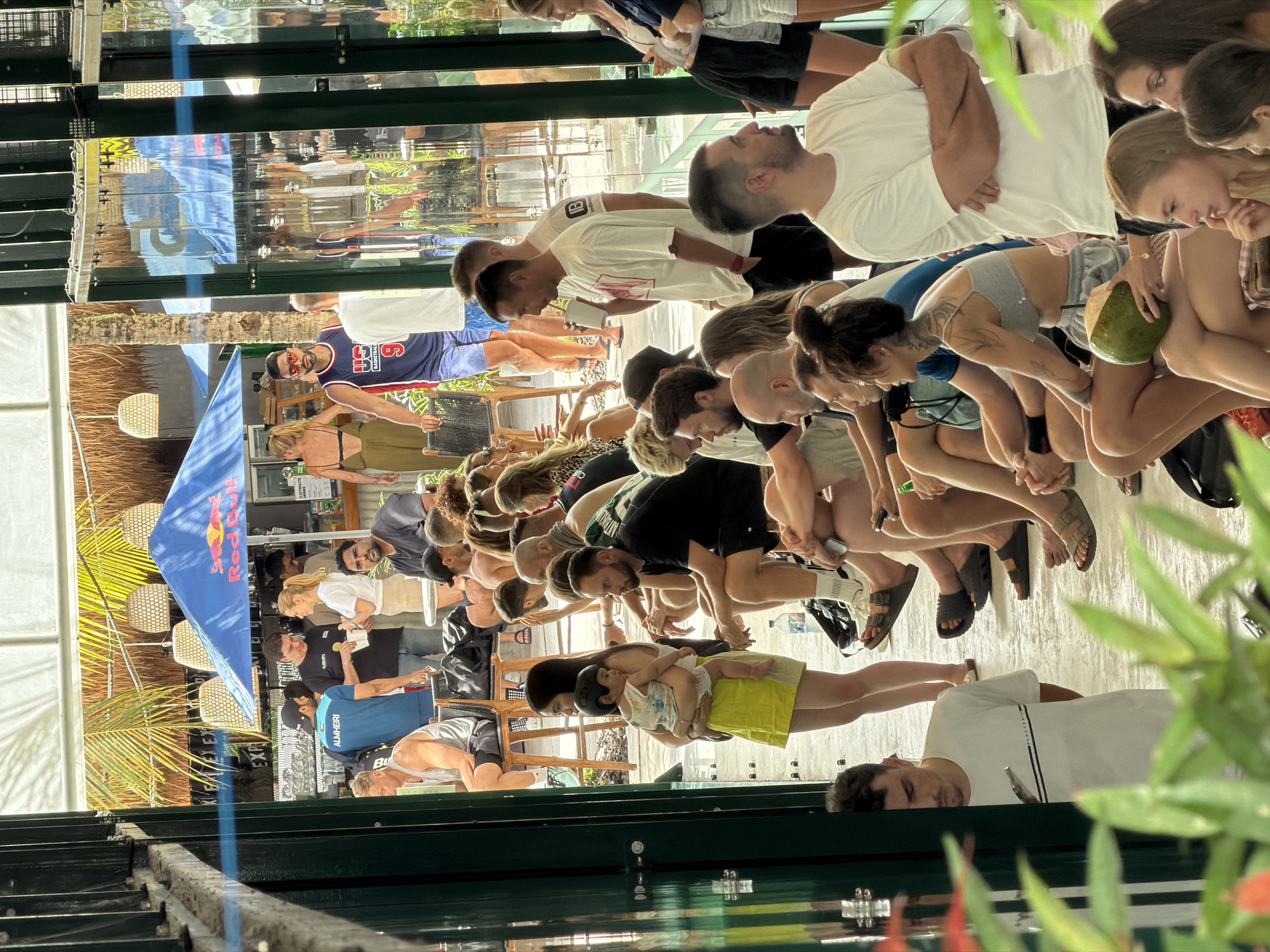 A photograph depicts a group of approximately 20 individuals, predominantly adults, in what appears to be an outdoor or semi-outdoor setting. The scene is illuminated by natural light, casting shadows on the ground. Several people are sitting on chairs, facing forward with their heads down. One person is holding a coconut. There is a woman holding a child with a black helmet and light patterned shirt. Some people are standing and interacting with others seated. A blue Red Bull branded flag is visible, along with palm fronds and thatched roofing elements. The scene appears to be a social gathering or event. The location is North Kuta, Indonesia.