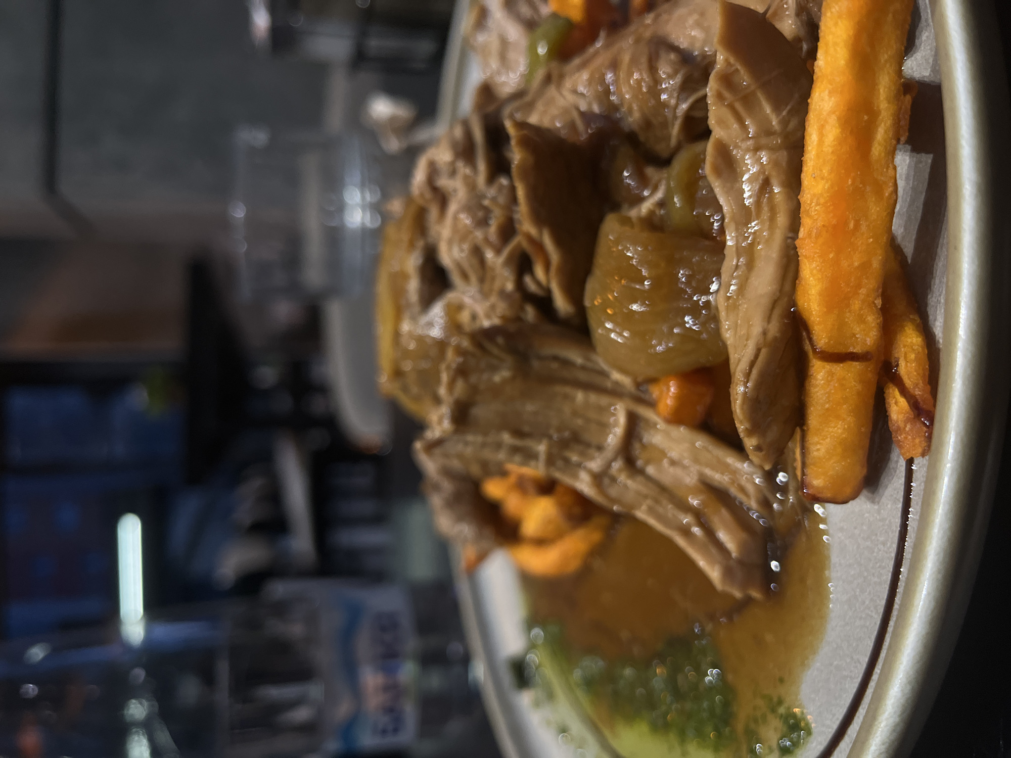 The image depicts a plate of food, likely served in a restaurant or dining establishment. The main components of the dish include braised or pulled meat, possibly pork or beef, along with sauteed vegetables such as peppers and carrots. Several thick-cut sweet potato fries are also included on the plate. A small pool of sauce or jus is present. The plate appears to be ceramic. The blurred background shows a suggestion of a restaurant interior. There is no specific indication of the location in Blagoevgrad, Bulgaria, however.