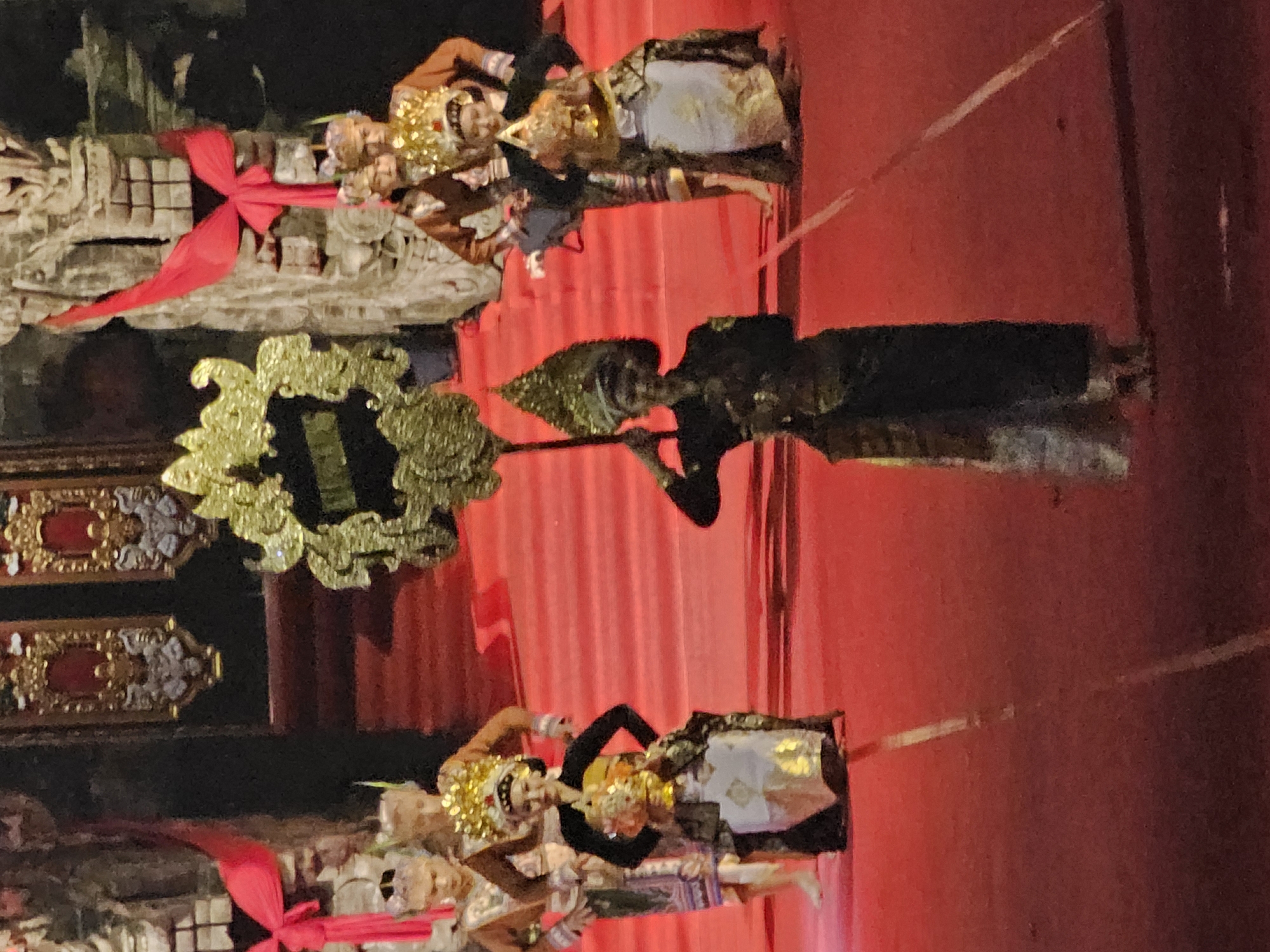 A group of people in traditional Balinese attire are performing on a red carpeted stage. The foreground features two individuals in elaborate costumes. The person on the right is holding a golden, ornate staff and wears a dark tunic over a patterned skirt, and a detailed headdress. The person to the left is dressed in a black top, a yellow and gold garment, and a white and gold skirt, also adorned with a headdress and holding a golden offering. Behind them, several other performers in similar traditional dress are visible. The background shows a dark structure with intricate carvings and red draped fabric. The overall scene suggests a cultural performance, possibly a dance or religious ceremony, taking place in Denpasar, Indonesia, given the context of traditional Balinese attire.