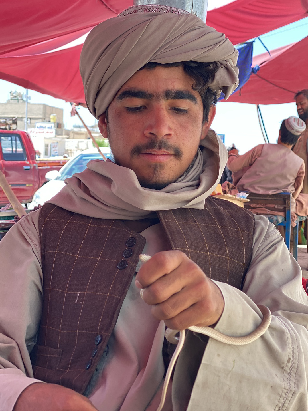 In Qandahar, Afghanistan, a man wearing a light brown turban and a checkered vest is holding a small, pale snake. He gazes downward at the reptile coiled in his hand. The scene is set outdoors, under a red canopy, with other people and vehicles visible in the background, suggesting a public place.
