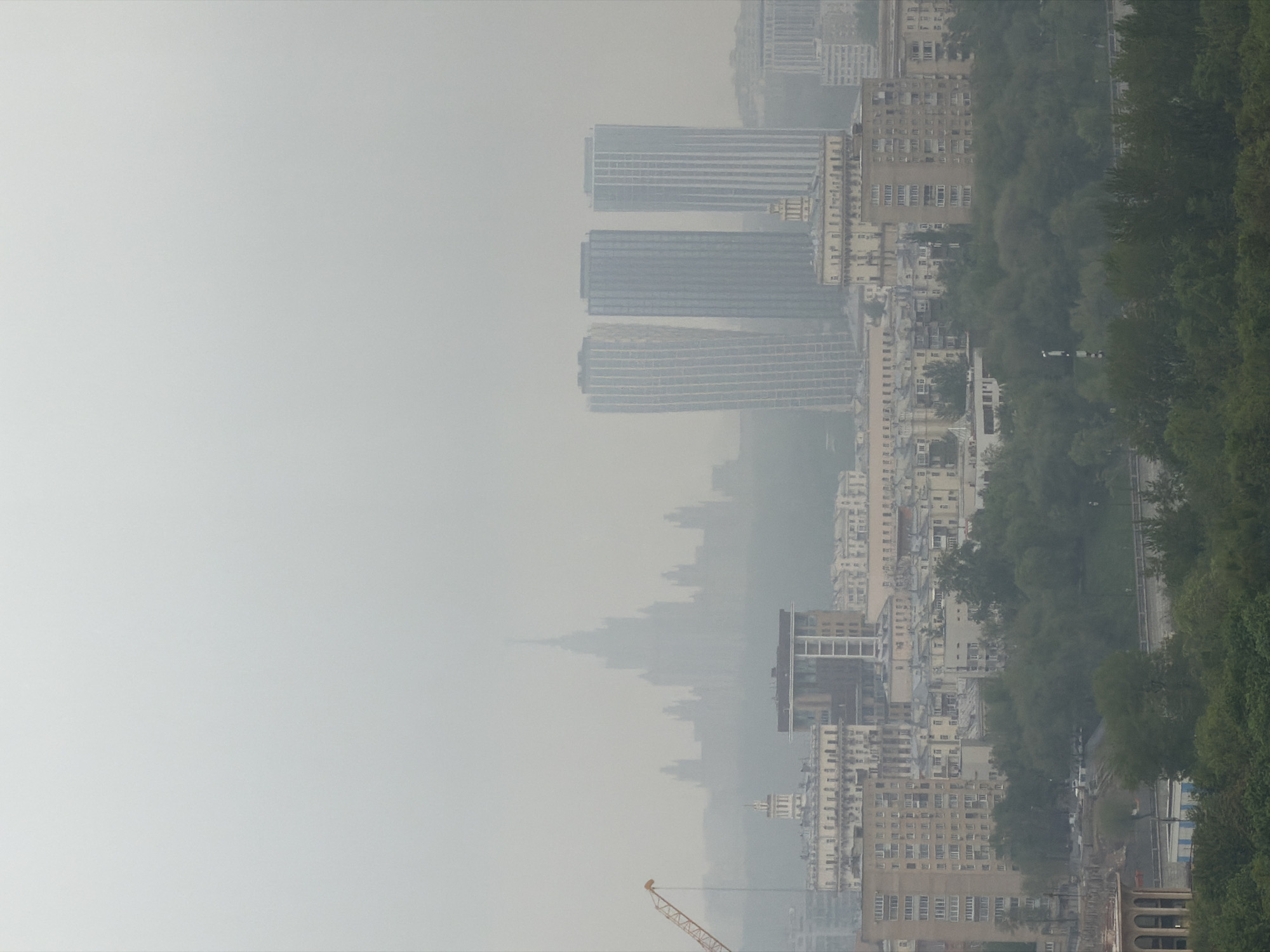 The image shows a cityscape in Moscow, Russia, heavily obscured by smog or fog. Notable are the high-rise buildings, including a cluster of three modern skyscrapers identifiable as part of the Moscow City business district. There are residential buildings with smaller stories and the surrounding green foliage indicate the urban environment blending with nature. The atmosphere has a strong haze that softens details. A construction crane is visible toward the bottom left corner.