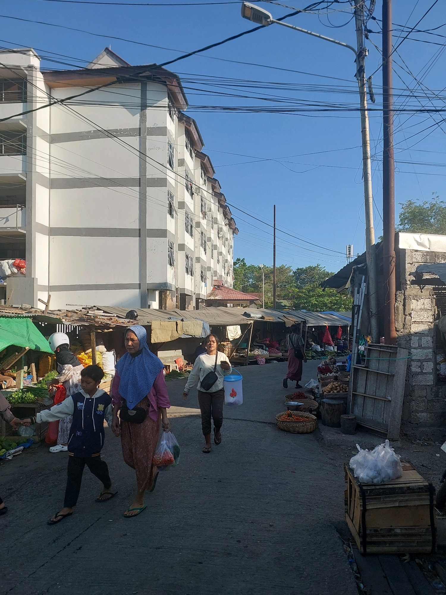 This is a medium-shot, eye-level, outdoor, daytime photograph of a street market in Kupang, Indonesia. The background features a large, multi-story building with a white facade and grey horizontal stripes. The building has numerous windows and balconies. In the foreground and midground, there are several market stalls with makeshift roofs constructed from wood, corrugated metal, and tarps. These stalls display various produce, including vegetables and fruits.
Several people are visible. In the midground, a woman wearing a blue hijab and a pink top, and a patterned pink skirt walks towards the viewer, holding a plastic bag of produce. Beside her, a young boy in a blue and white jacket and dark pants walks with his left hand held by the woman. Another woman, wearing a light-colored long-sleeved shirt and dark pants, walks away from the viewer towards the right side of the frame. Further in the background, other individuals are seen browsing or working at the market stalls.
The street is paved with asphalt. Overhead, a complex network of electrical wires and a street lamp are visible against a clear blue sky. Several utility poles are also present, one of which is significantly thicker and darker than the others. In the right foreground, a wooden crate is visible, topped with a pile of white plastic bags. The overall impression is that of a busy, functioning open-air market in an urban setting.