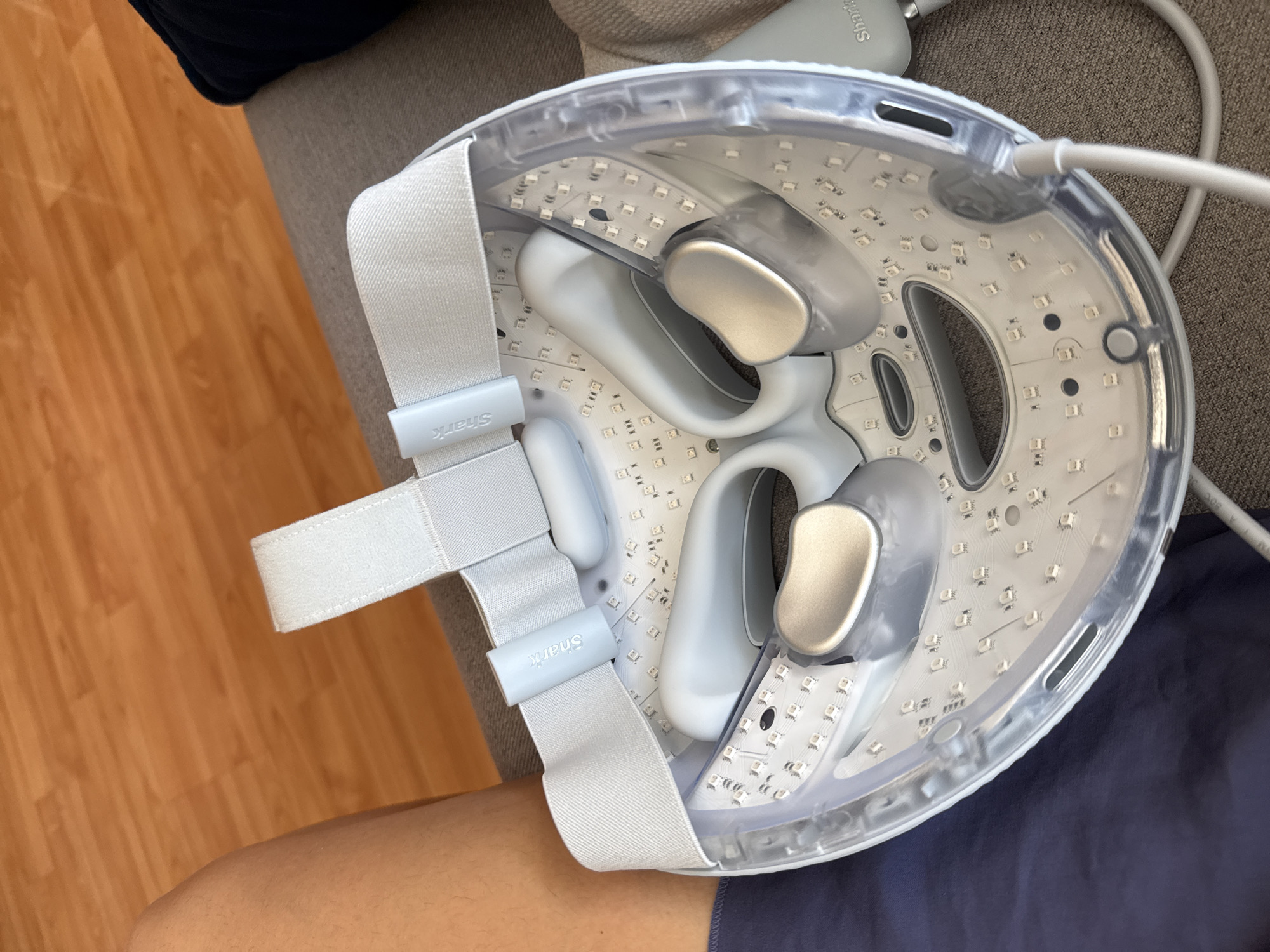 The image captures an indoor scene, likely a home, showing a close-up of a light therapy face mask or similar skincare device. The device is resting on a person's lap, which is partially visible at the bottom of the frame, clad in dark blue or navy fabric.
The primary subject is the white and translucent face mask, designed with numerous small, square LED lights arranged across its interior surface. It features soft, grey padding for the eye and nose areas, suggesting comfort during use. Adjustable light grey fabric straps with Velcro fasteners are attached to the mask, intended for securing it to a user's head. Two light grey plastic adjusters on these straps clearly display the brand name "Shark" in white text. Another grey cylindrical component, possibly part of the power supply or control unit, also has the "Shark" logo visible on its side, located at the upper right. A grey cable extends from the mask, disappearing out of frame.
The device is positioned against a background that includes a beige or light grey upholstered surface, likely a couch or chair, and a warm-toned wooden floor with visible grain. The lighting suggests an indoor environment, possibly daytime. No activity beyond the resting position of the device is depicted; the LED lights are not illuminated.