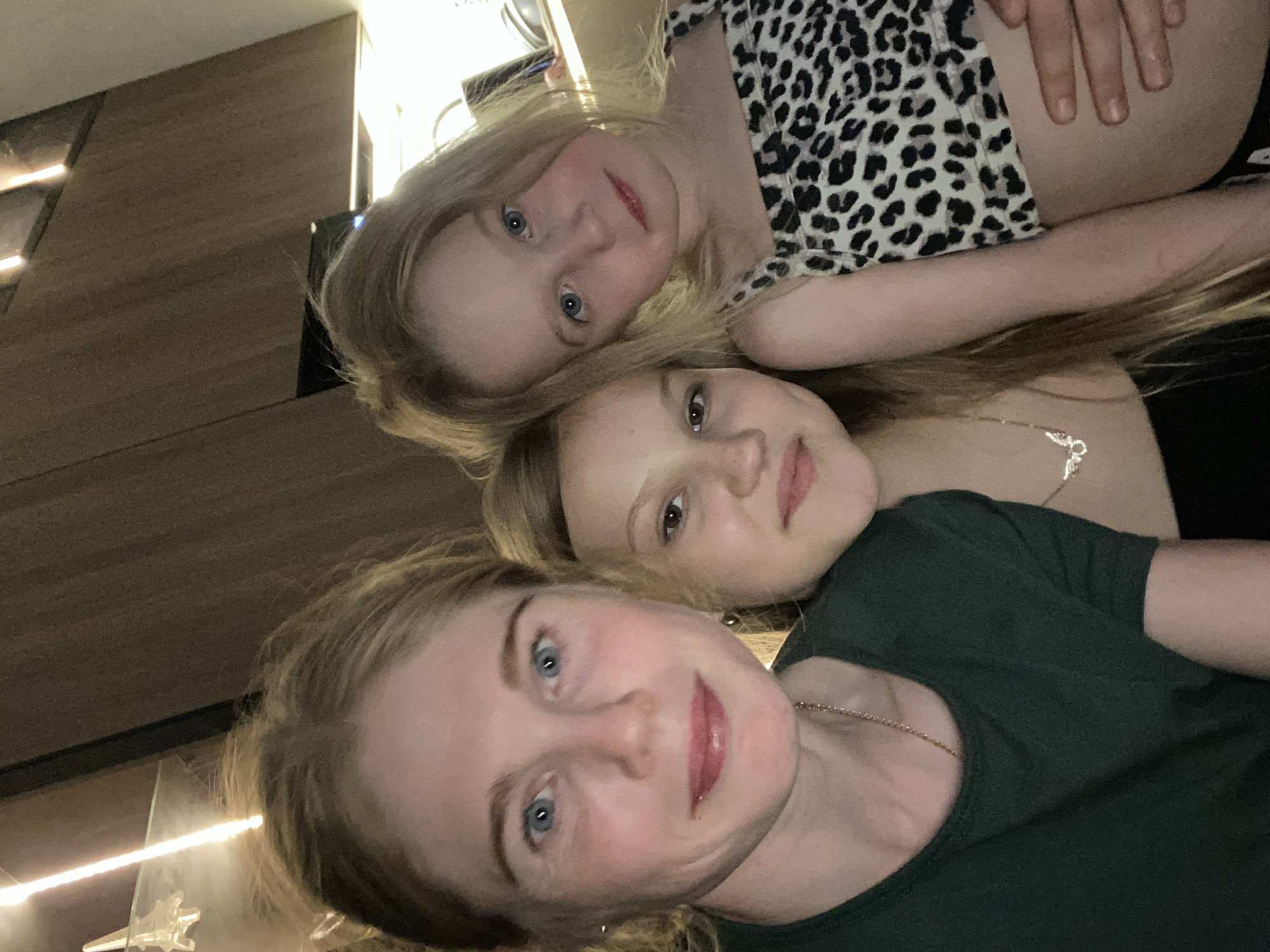 Three individuals are captured in a selfie-style image. At the bottom of the frame is a fair-skinned adult female with blue eyes and blonde hair, wearing a dark green shirt and a delicate gold necklace. Above her is a younger fair-skinned female, also with blonde hair, wearing a dark-colored shirt and a silver-colored necklace with a pendant. At the top of the image is a young fair-skinned female child with blonde hair and blue eyes, wearing a spotted top with a light-colored background. A hand rests on the child's stomach. The setting appears to be indoors, with a dark background and a wooden paneled wall partially visible. No specific landmarks identify the location as Chaikovsky, Russia.
