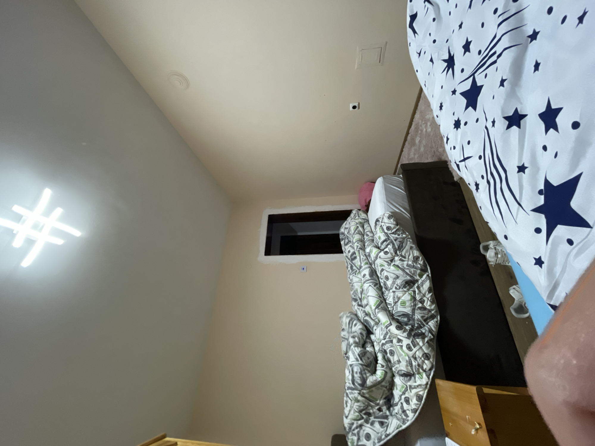 A dimly lit interior space, possibly a bedroom, is observed. The primary focus is a partially visible bed covered with a patterned comforter featuring blue stars on a white background. A second comforter with a money pattern drapes over the side of the bed. A dark window is visible against a beige wall, and there appears to be a pink pillow on the bed. The room is not identifiable as being in Presoka, Bulgaria, based on the information available in the image.
