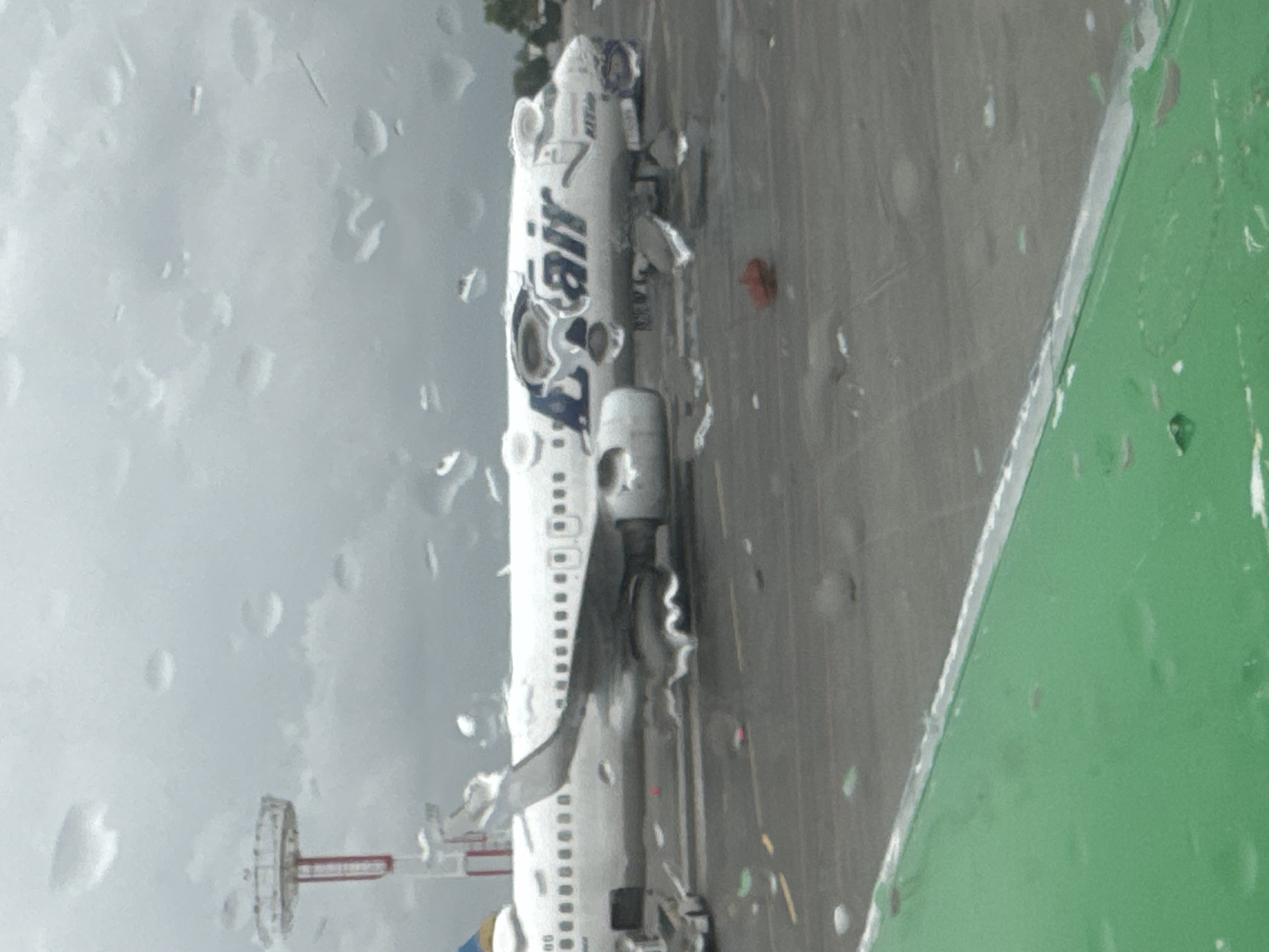 The image shows an "Uzbekistan Airways" airplane parked on an airport tarmac during a rainy day. The scene is viewed through a raindrop-covered window. In the background, a control tower and other airport infrastructure are visible. The location is likely Tashkent, Uzbekistan, given the airline's name.