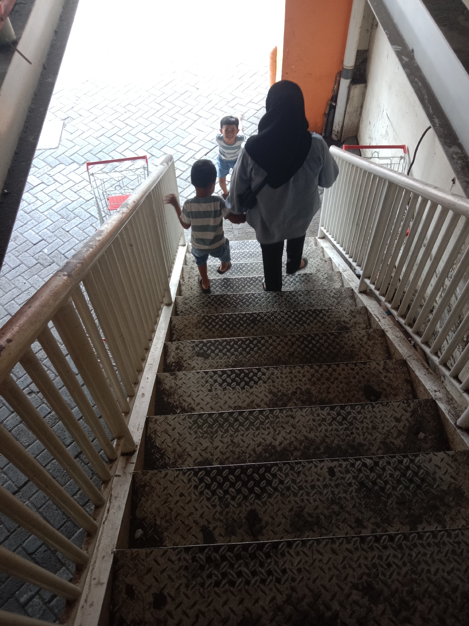 The image shows a person in a black hijab and a grey shirt, followed by two young boys walking down a metal staircase. The boy in the front is wearing a striped t-shirt and shorts, while the boy behind him is wearing a blue and white striped t-shirt and shorts. The staircase is outdoors, with a light-colored railing on the left. Below the staircase, there is a paved area with interlocking bricks. Two shopping carts are visible on the paved area, one red and one silver. The surroundings suggest a shopping area or mall entrance. This scene is located in Probolinggo, Indonesia.