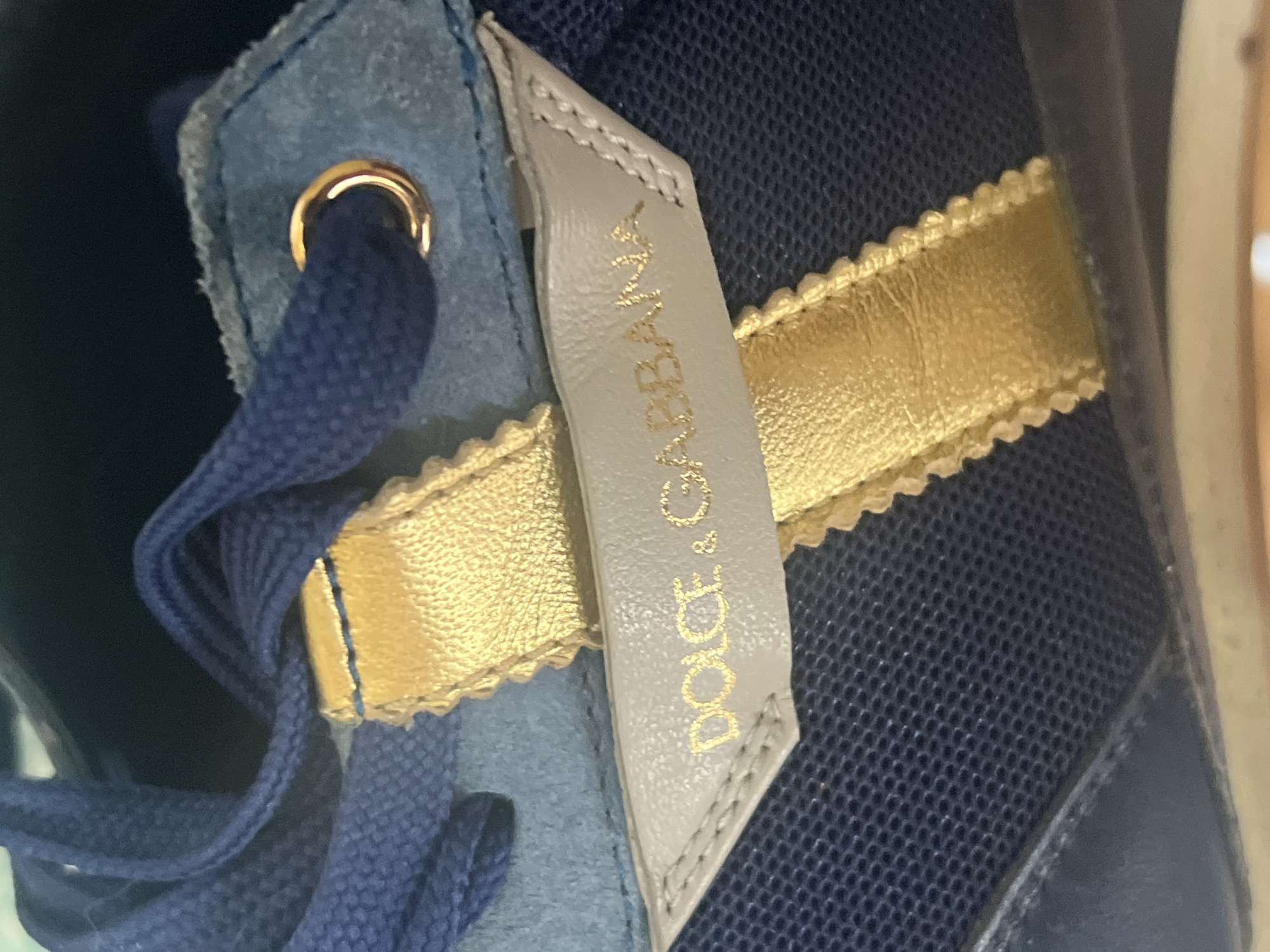This is a close-up, detailed image of a blue Dolce & Gabbana sneaker. The shoe features blue laces, a light blue suede accent, and a tan leather tag with gold lettering that reads "DOLCE & GABBANA." A golden ribbon detail is attached to the tag. The sneaker is made of a dark blue mesh material and a darker blue leather or synthetic material. The image does not contain any information about the city of Sochi, Russia, or any people or actions.