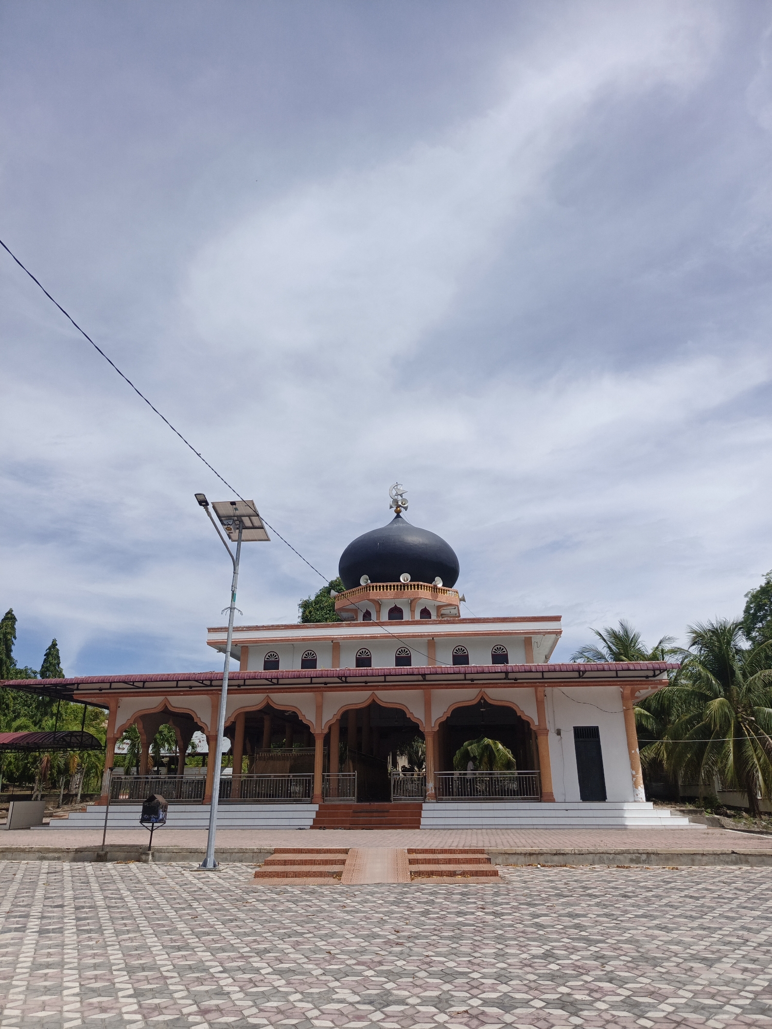 The image shows a mosque located in the city of Sawang, Indonesia. The mosque has a large black dome and is surrounded by palm trees. The building is white with orange trim and features arched walkways. In front of the mosque, there is a paved courtyard with a solar-powered street light. The sky is partly cloudy. There are no people visible in the image.