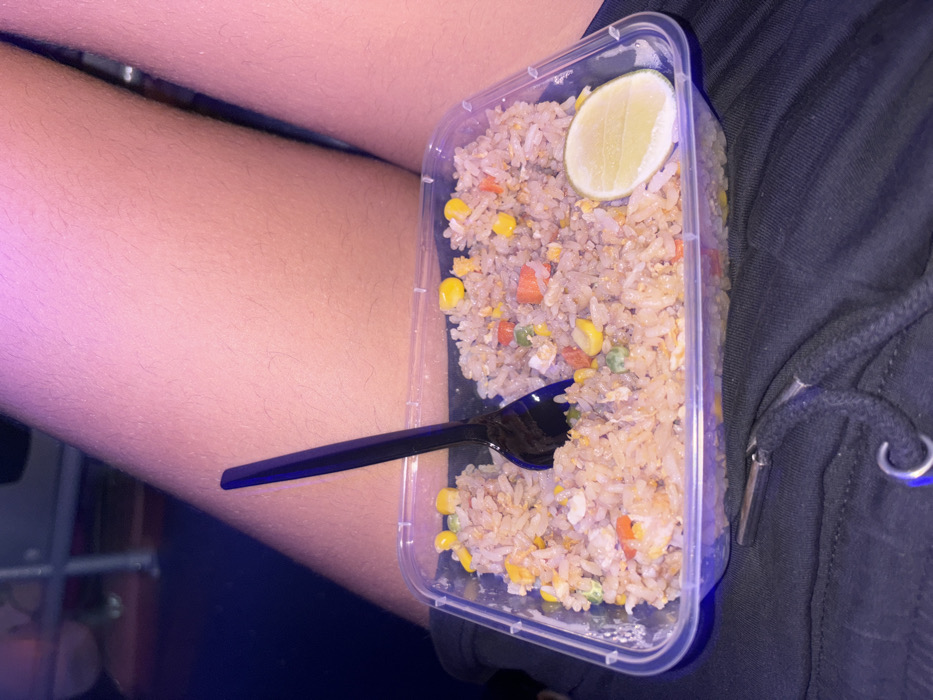 The image displays a clear plastic rectangular takeaway container filled with fried rice, positioned on dark-colored fabric, likely shorts or pants. The fried rice consists of white rice grains, yellow corn kernels, green peas, and diced orange carrots, with visible specks of scrambled egg. A black plastic spoon is partially submerged in the center of the rice, its handle extending upwards. A light green lime wedge rests on the right side of the rice. The dark fabric below the container features a black drawstring with silver-colored metal aglets and a silver-colored metal ring. Above the container, portions of a person's bare upper thighs are visible, exhibiting fair skin with fine hair, illuminated by a strong pinkish-purple artificial light source. The background is dark and out of focus, with an indistinct reflective or metallic element visible in the upper left. The exact location within Surat Thani City Municipality, Thailand, is not identifiable from the visual content.