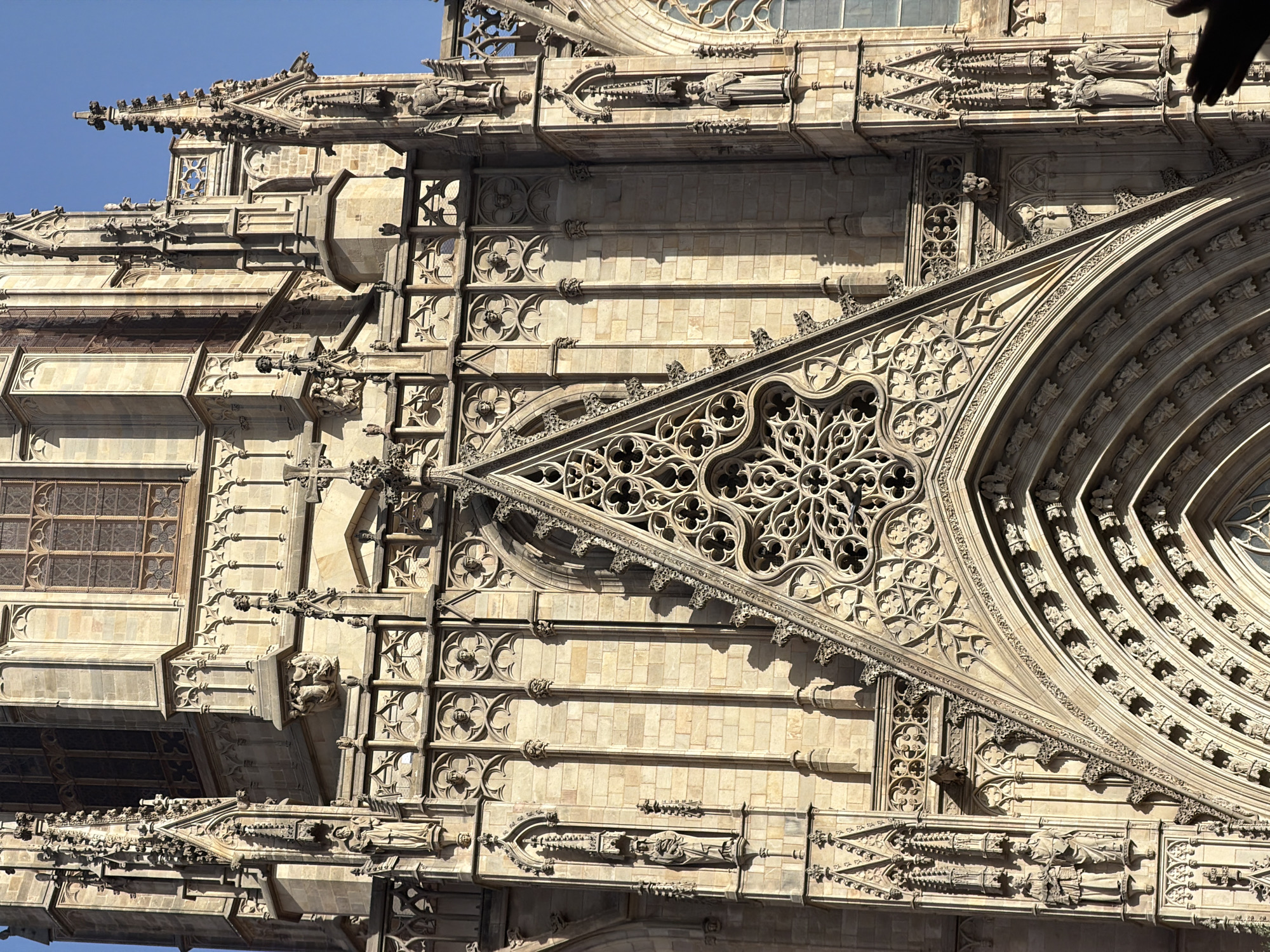 The image shows a close-up, frontal view of the facade of the Barcelona Cathedral, a Gothic cathedral located in Barcelona, Spain. The architecture is characterized by intricate stonework, including pointed arches, flying buttresses, and detailed sculptures. In the center of the image is a large, triangular pediment filled with delicate tracery, a common feature of Gothic architecture. Below this, a series of arched doorways are visible, adorned with decorative carvings. Small statues of figures, likely saints or biblical characters, are set into niches along the facade. The stone of the building appears weathered and aged, with variations in color due to sunlight and shadow. The sky above is a clear, bright blue. The overall impression is one of grand, historical religious architecture.