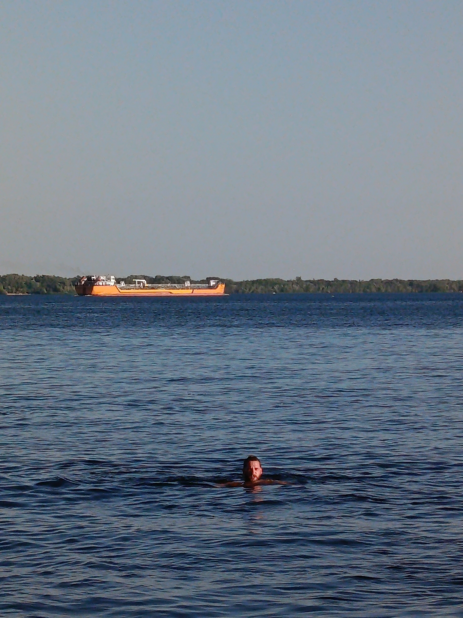 A man is swimming in a body of water. He is facing the camera with his head and shoulders visible above the water. In the background, a large orange ship is sailing on the water. A treeline is visible on the horizon behind the ship. The sky is clear and pale blue.