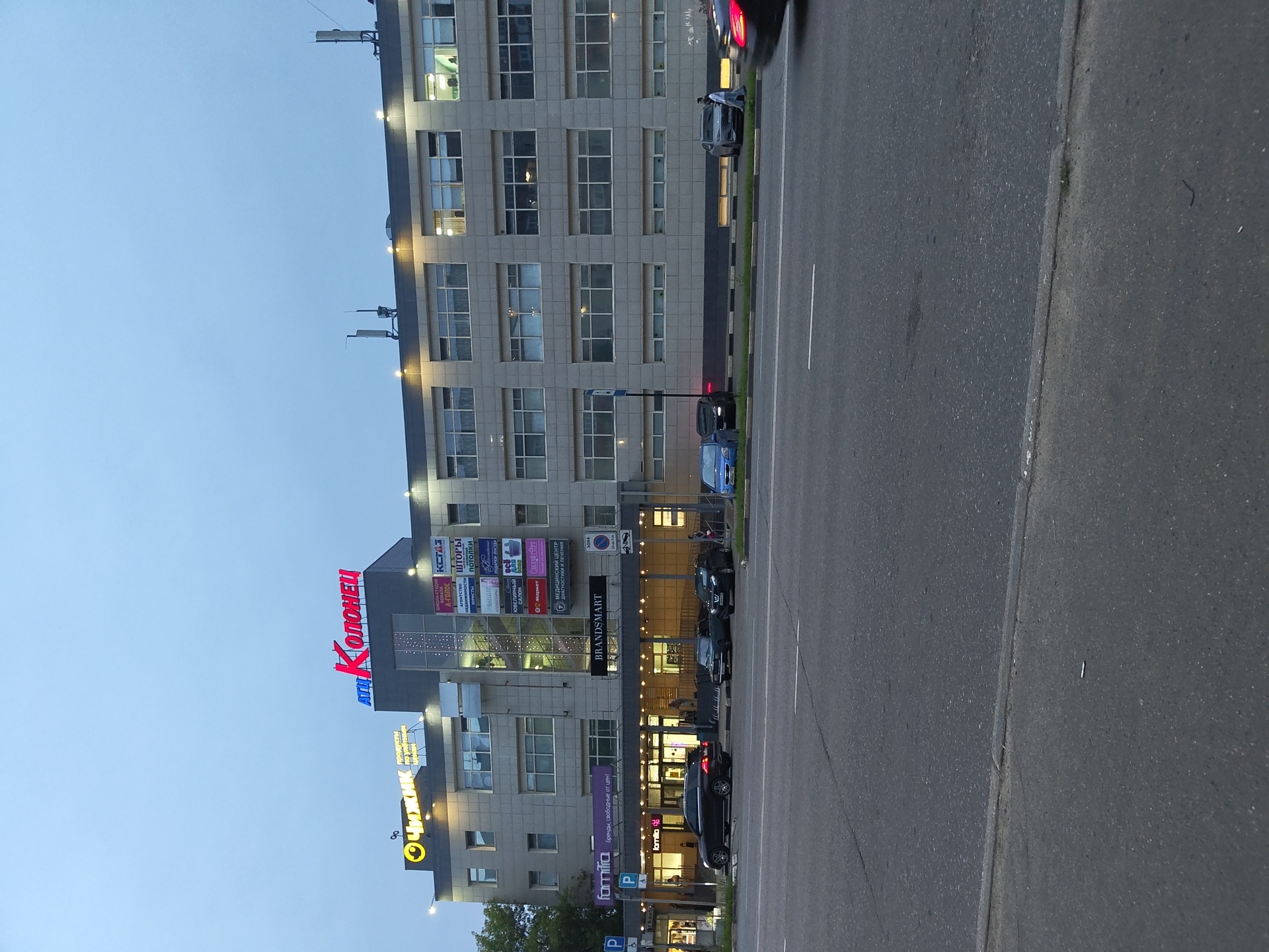 The image shows a multistory commercial building with a sign reading "АТЦ КЛ ОЛОНЕЦ" at the top. Below this, there are various smaller signs indicating shops and services, including "Чижик" (a grocery store), "BRANDS MART," "familia" (a clothing store), "А-ГОЛОС," "ШТОРЫ," "КСТАЗ," and "МЕДИЦИНСКИЙ ЦЕНТР ДИАГНОСТИКИ И ЛЕЧЕНИЯ." The building is situated on a street with several cars parked along the sidewalk and a road with traffic in the foreground. The sky is overcast and appears to be twilight. The architecture suggests a modern commercial district. This location is identified as the ATs Kolonets in Zhukovsky, Russia. There are no individuals clearly visible in the act of interaction, and the scene appears to be a street view of a commercial establishment.