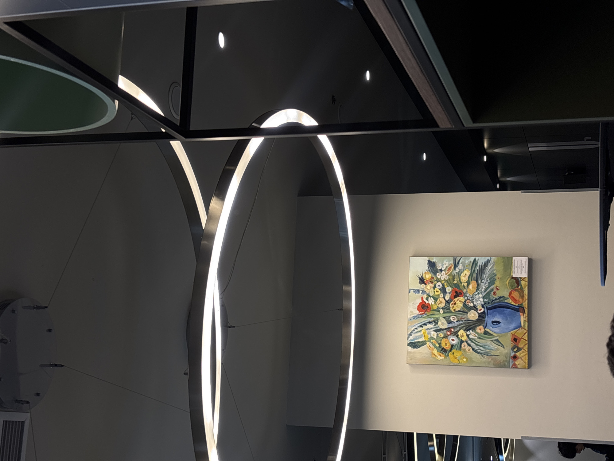 The image depicts an interior scene. The foreground features two prominent, vertically oriented, circular light fixtures with integrated LED strips. The fixtures are silver-colored. Behind these, a wall is visible, upon which hangs a painting of flowers in a vase. The painting is set against a light-colored wall. The ceiling above is dark and reflective, with several small, circular lights embedded within it. The composition is somewhat disorienting, potentially due to reflections and unusual angles. The exact location in Yerevan, Armenia, is undeterminable without further information. No people are wholly visible in the image. There are elements of a person at the bottom right of the frame.