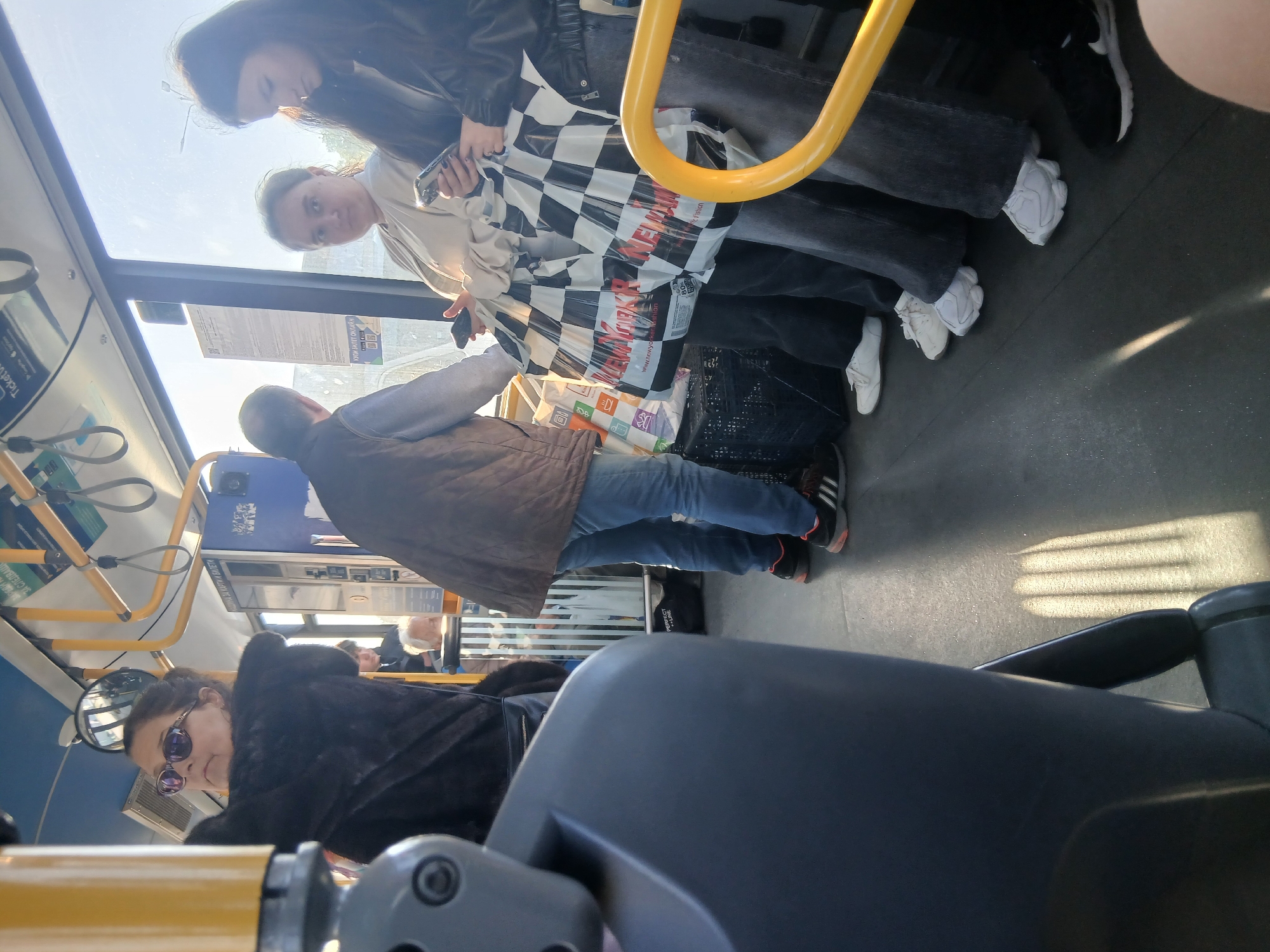 The image depicts the interior of a public transportation vehicle, likely a bus, in what appears to be Varna, Bulgaria, based on visual cues. The scene includes a group of several people. Two women are standing near a yellow handrail. One is looking at a phone and holding a shopping bag with a checkered pattern. Behind her is a woman in a leather jacket. Nearer the foreground, a man in a brown vest and jeans stands, also holding bags. Further back, other seated or standing passengers are visible. The foreground is dominated by a bus seat, which is slightly out of focus. The lighting is bright, suggesting a sunny day. The scene shows everyday life on public transport. A machine for cards and tickets is also visible.