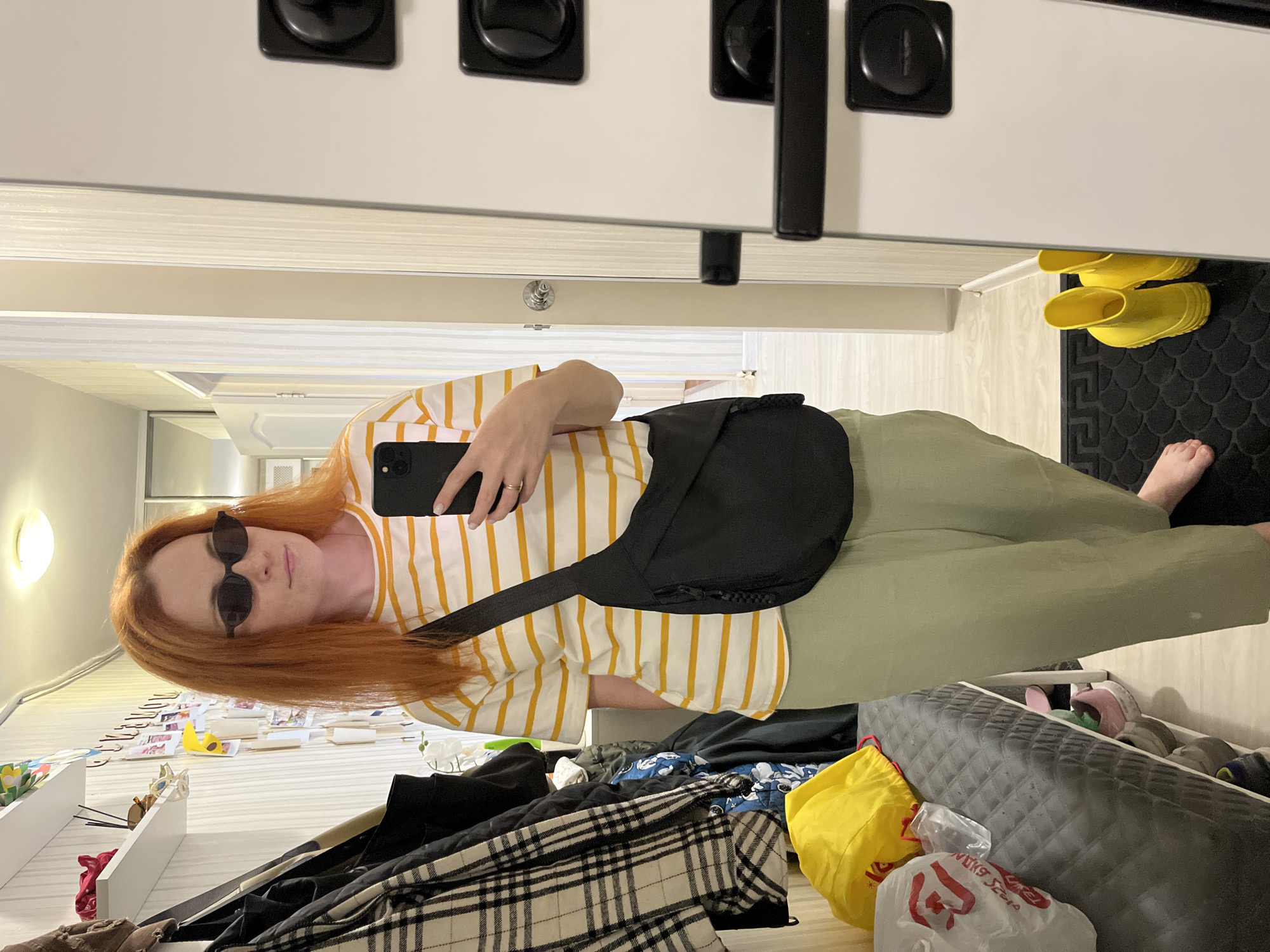 The image, taken in Chaikovsky, Russia, is a full-length, vertically oriented mirror selfie of a young woman with auburn hair. She is wearing a yellow and white striped t-shirt, light green pants, and black sunglasses. A black crossbody bag is slung over her shoulder. Her left hand holds a black smartphone, with which she is taking the photo. A ring is visible on her left ring finger.
The background suggests an indoor setting, possibly a hallway or dressing area. To the left of the woman, a wall with several black square light switches or electrical outlets is visible. To the right, a pair of bright yellow rain boots is placed on a black mat. Below this, a glimpse of a floor with a wood-grain pattern is seen.
In the reflection, below the woman, various items are visible. There are shelves filled with what appear to be photographs or cards, a yellow banana-shaped toy, and some white flowers. Further to the right, a cluttered area includes clothing items like a black jacket and a black and white plaid garment, along with a yellow bag and a white plastic bag with red lettering that is partially obscured but appears to say "Viking Store". The overall impression is of a casual, personal snapshot, likely taken in the woman's home. The lighting suggests it is daytime, and there are no indications of specific weather conditions. The woman's expression is neutral.