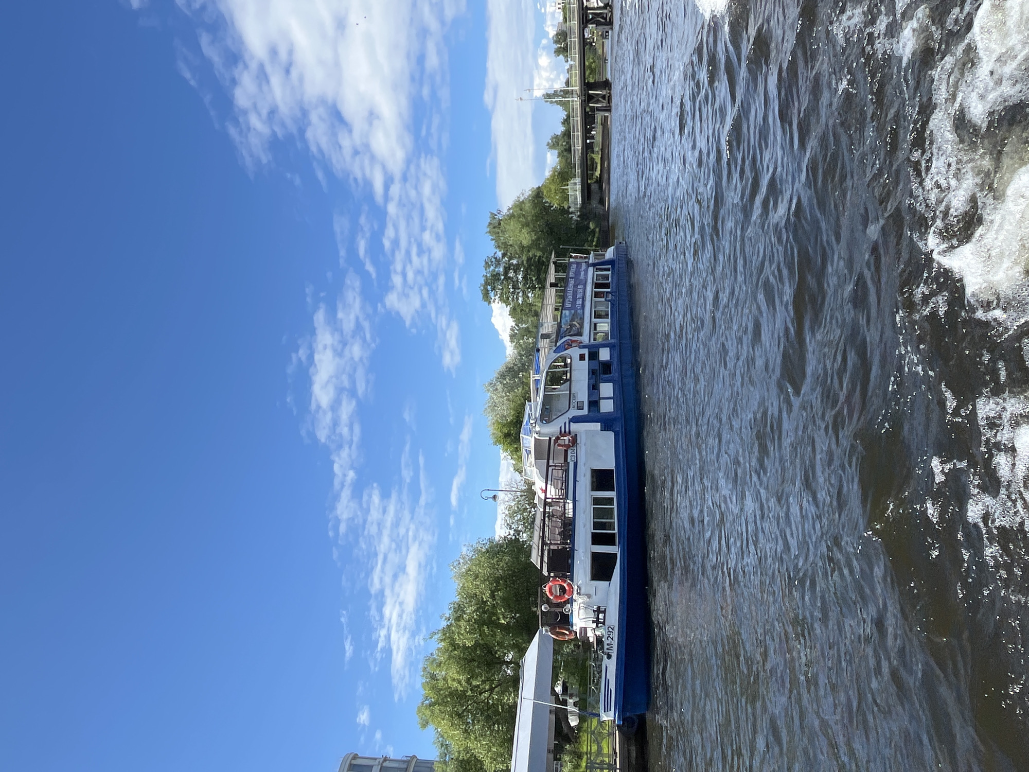 This media file shows a blue and white passenger boat docked on a river in Saint Petersburg, Russia. The boat has multiple windows and a covered upper deck with seating. Several life preservers are visible on the boat's exterior. Trees and greenery line the riverbank, and a bridge structure is visible in the distance. The sky is blue with scattered white clouds, suggesting a pleasant, sunny day. The water is rippling, indicating gentle movement. The scene appears calm and peaceful, possibly during a leisurely boat tour or as the boat awaits passengers. Text on the boat's side is partially visible, reading "M-292" and some Cyrillic characters, likely indicating the boat's designation or route. The overall impression is a typical summer day in a scenic riverside location.
