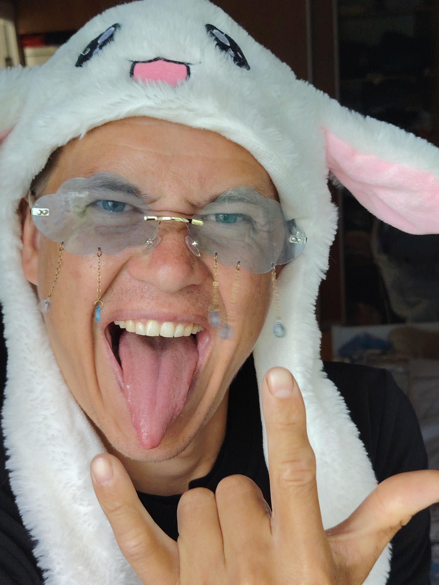 A close-up, eye-level shot shows a person wearing a white plush bunny hat with pink ear lining and black eyes and nose details. They are also wearing unique, rimless, cloud-shaped glasses with gold accents. Dangling from the glasses are delicate gold chains with small blue and clear beads. The person's mouth is open wide, with their tongue extended, and their teeth are visible. Their right hand is raised, with the index and pinky fingers extended in a "rock on" gesture. The background is blurred, but appears to be indoors, possibly a room with dark shelving or furniture. The image does not provide any information or context related to Chaikovsky, Russia.