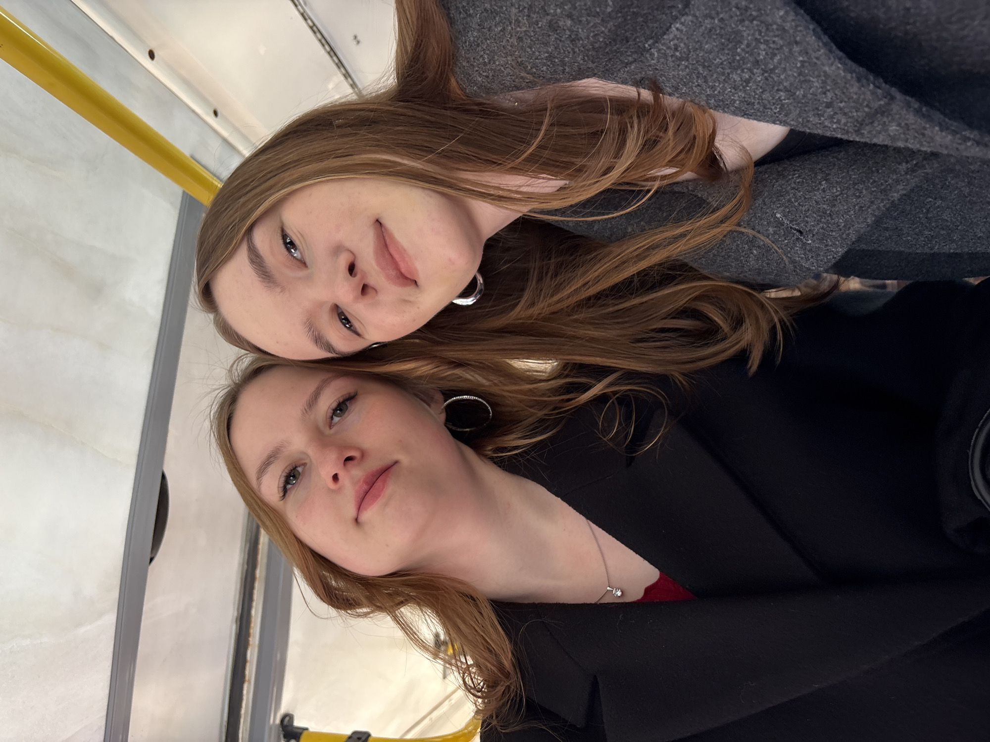 Two young women pose for a selfie on public transportation in Kazan, Russia. The upper woman has light brown hair and is wearing a gray coat. The lower woman has light brown hair and is wearing a black coat. Both women are wearing hoop earrings. The background shows a yellow handrail and the interior of the bus or train.