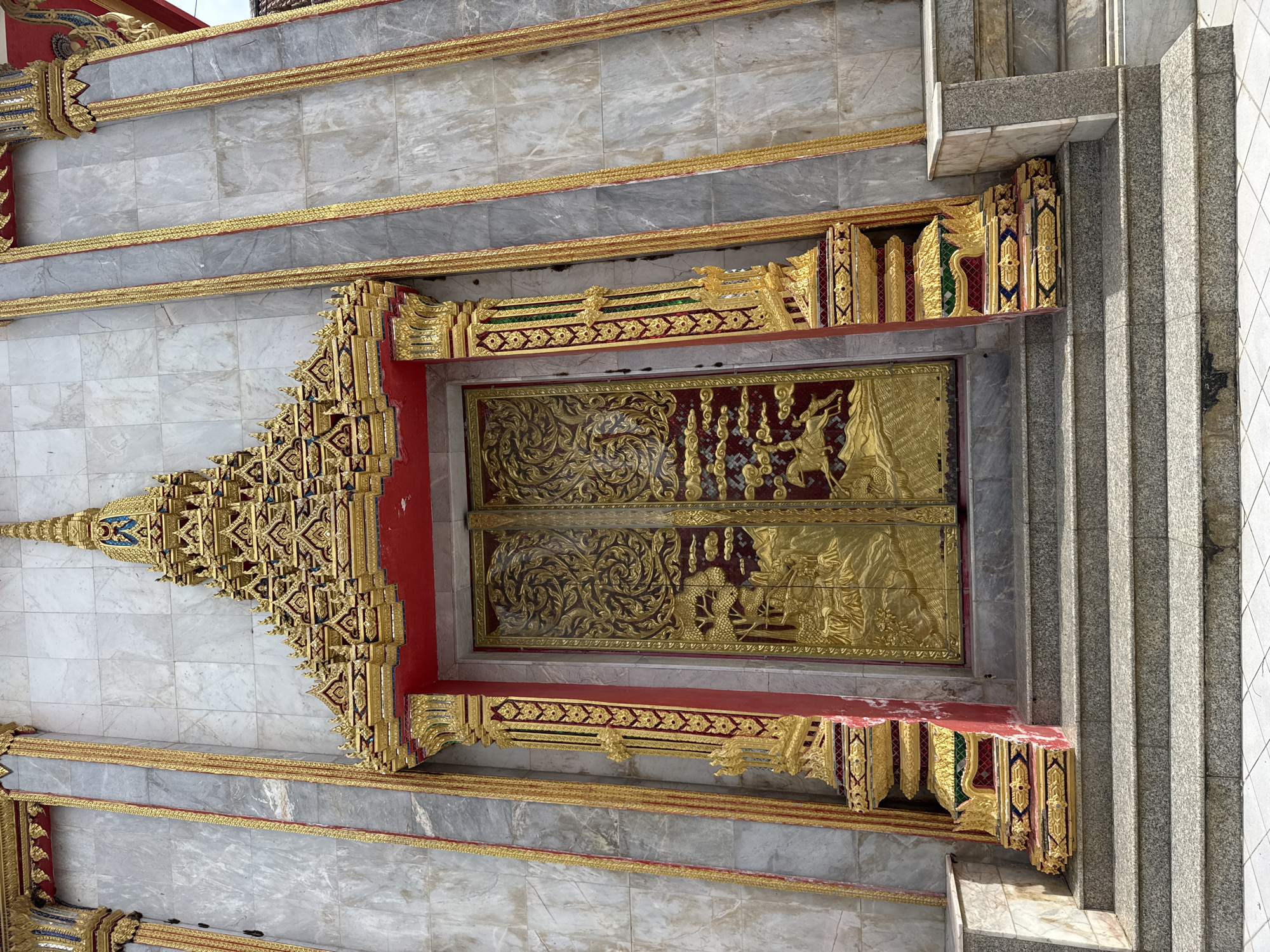 This image shows a close-up, eye-level view of the ornate entrance to a Buddhist temple in Chalong, Thailand. The focal point is a grand set of double doors, intricately carved and gilded with scenes of what appear to be mythological figures and landscapes. The doors are framed by richly decorated golden pillars and an elaborate, multi-tiered golden roof structure adorned with red accents and small spires. The walls surrounding the entrance are clad in large, polished grey marble tiles, segmented by vertical golden decorative bands. A set of grey granite stairs with matching granite risers leads up to the entrance. The lower section of the entrance features ornate golden and colored decorative bases, matching the style of the pillars and roof. The overall impression is one of opulence and detailed craftsmanship, characteristic of Thai temple architecture.