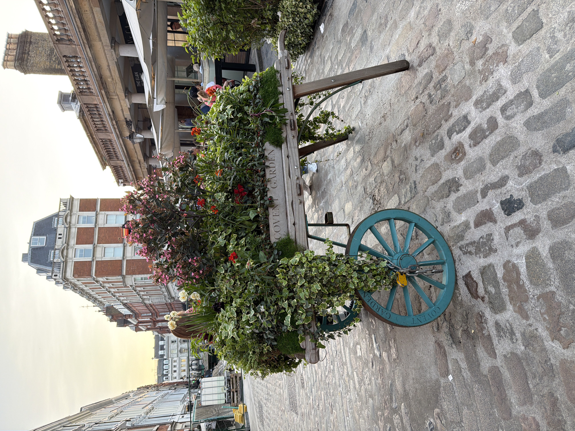 The image shows a flower-filled cart in Covent Garden, London, United Kingdom. The cart, painted a muted blue-green, is overflowing with a variety of plants and flowers in shades of green, red, pink, and white. The words "Covent Garden" are visible on the side of the wheel. The cart is parked on a cobblestone street. Behind the cart, various buildings and shops are visible, including a multi-story brick building and a covered arcade with columns.