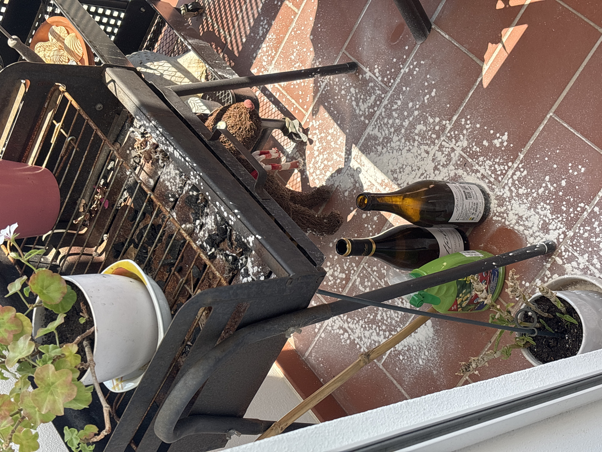 An outdoor balcony in Tavira, Portugal, features a charcoal grill on a metal cart, scattered with ash and debris. A potted plant with green and pinkish leaves sits on the grill's upper rack. Two wine bottles and a green spray bottle are placed on the tiled floor, which is covered in a fine white powder. A dried plant in a white pot is located near the edge of the balcony, next to a bamboo pole and a support for the grill. A decorative brown item resembling a bird is attached to the grill's structure. Sunlight casts shadows across the scene.