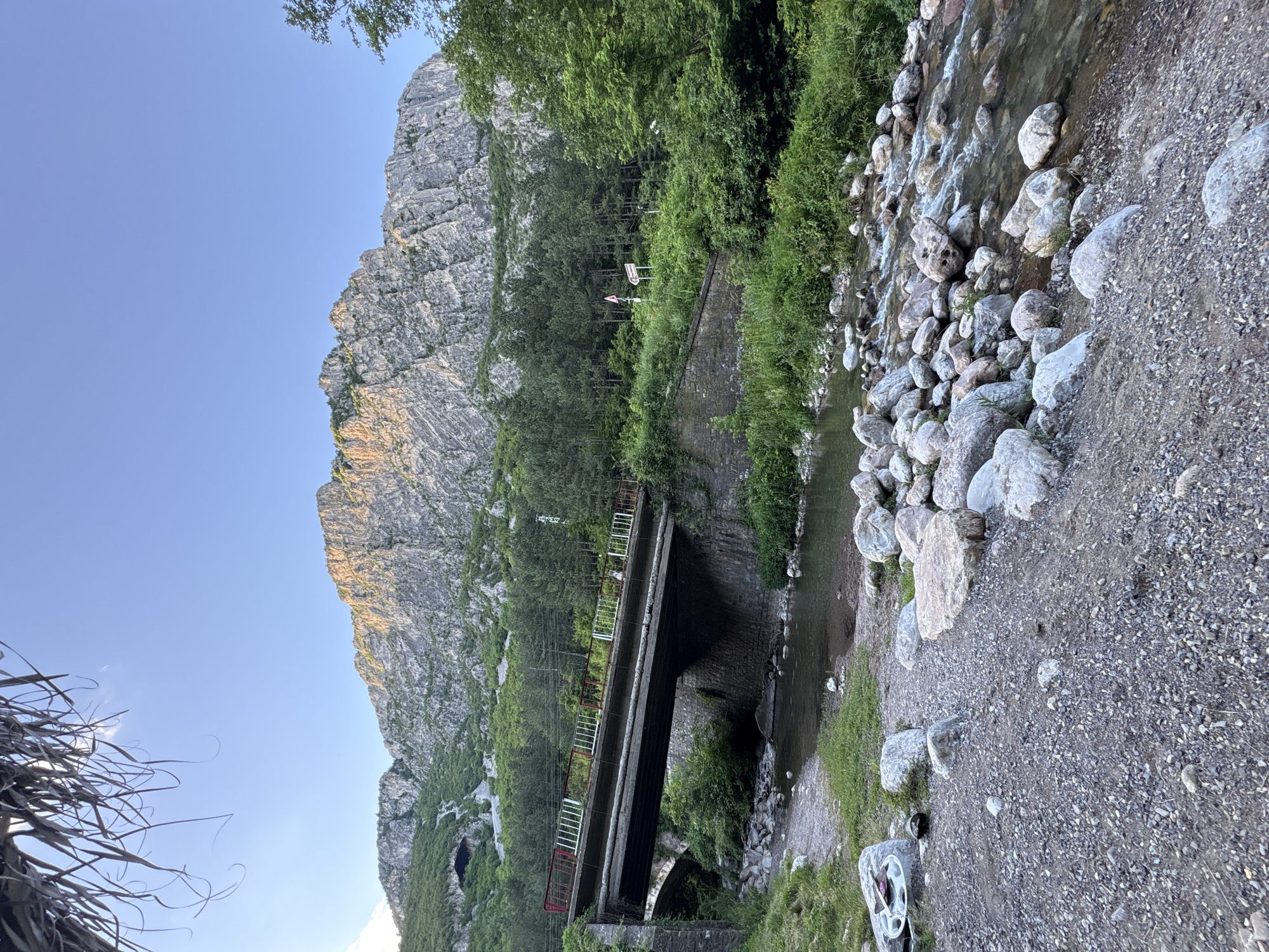 The image depicts a scenic view of a rocky mountain landscape in Zgorigrad, Bulgaria. In the foreground, a shallow, clear river flows over a bed of smooth, light-colored stones. The riverbank is covered in gravel and scattered rocks, with patches of green grass. A concrete bridge, supported by stone abutments, spans the river. The bridge has weathered concrete railings with red and white accents. Behind the bridge, dense green trees form a lush forest that climbs the mountainside. The towering mountain in the background is rugged and rocky, with sunlight illuminating its textured surface. A dark opening, possibly a cave entrance, is visible on the left side of the mountain. The sky is a clear, pale blue, indicative of a bright, sunny day. The overall scene is tranquil and natural, showcasing the beauty of the Bulgarian countryside.