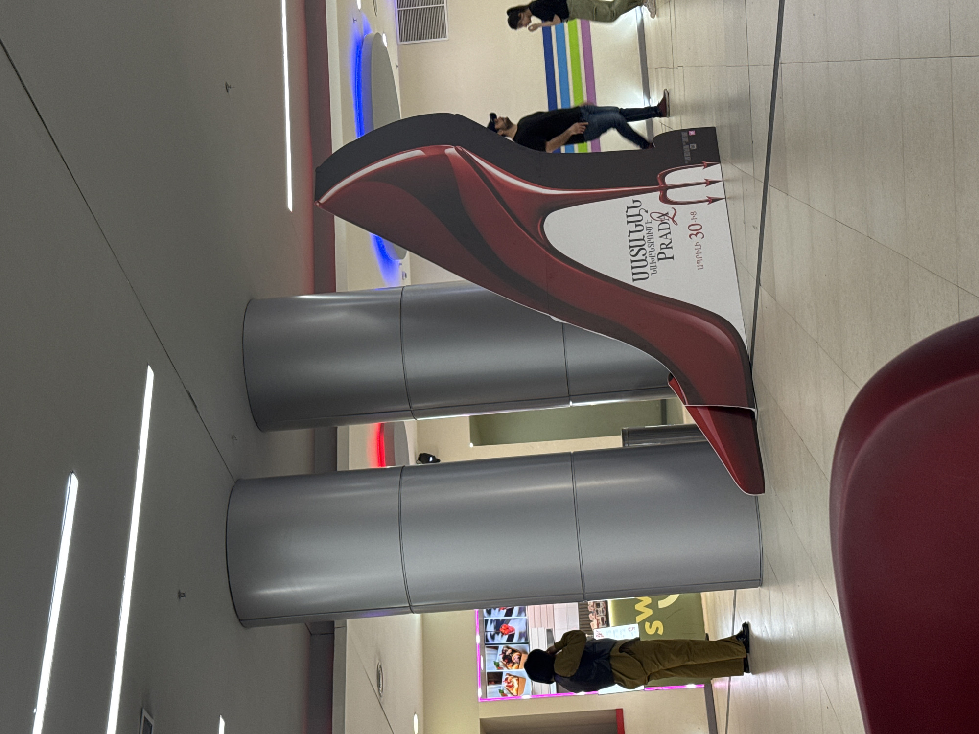 The photo depicts an indoor scene in Yerevan, Armenia, likely in a shopping mall or commercial building. There are two large cylindrical columns painted silver. Adjacent to them is a promotional cutout in the shape of a red, high-heeled shoe with a devil's tail. Above the shoe, text in Armenian script is visible, along with the word "PRADA." A man is lying on the floor, appearing to pose with the shoe cutout. Another individual wearing a green outfit and face mask walks in the background. Another person in khakis walks past a sign. The flooring is tiled in neutral tones. The ceiling features recessed linear lighting. The angle of the shot suggests the photograph was taken in an interior space.