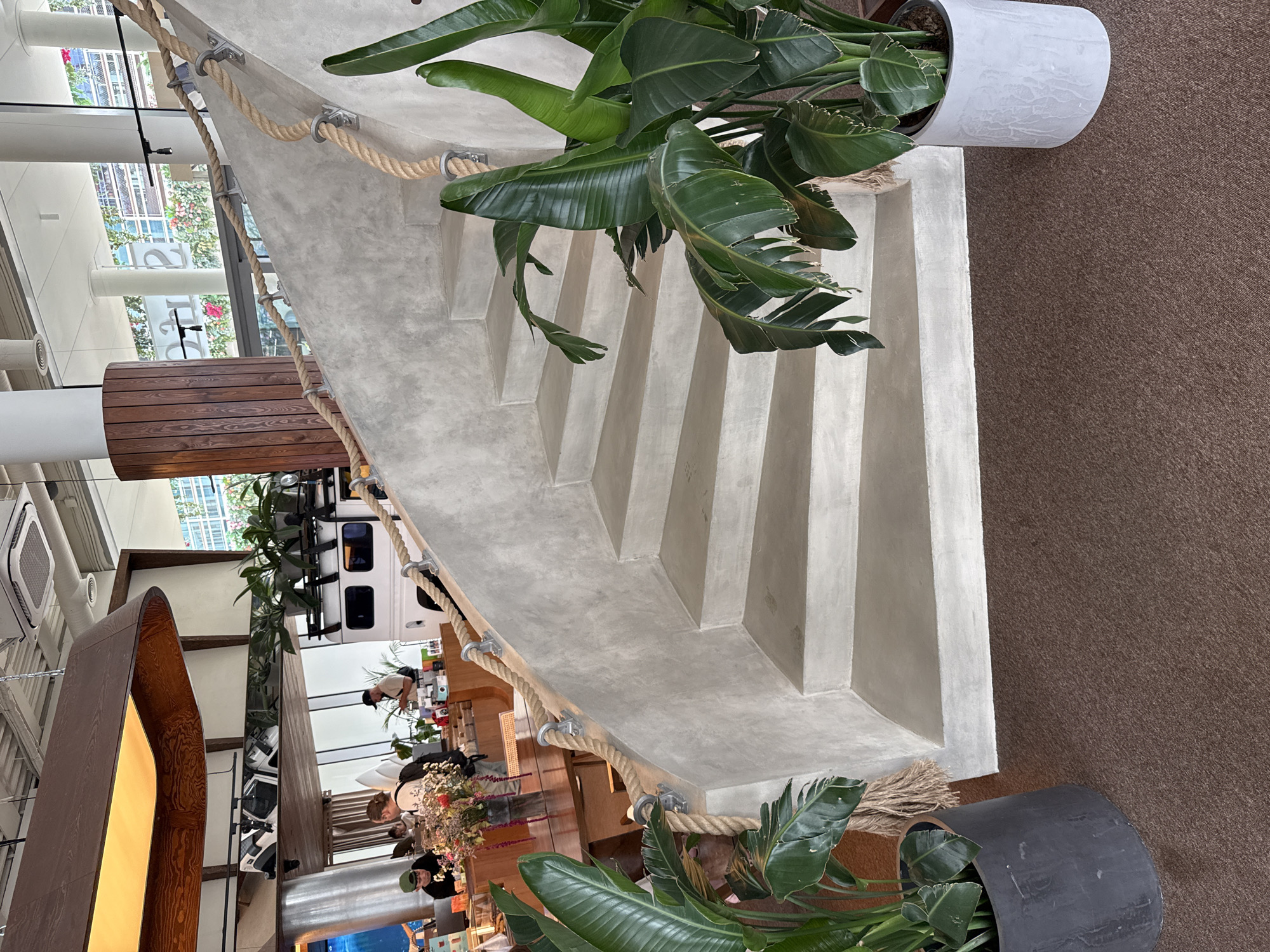 A concrete staircase with a rope railing is featured prominently in the foreground of this image, set within an indoor environment. The staircase curves upwards to the right, and the rope railing is secured by metal brackets to the concrete wall. Large green plants in dark gray and white planters are positioned at the base of the staircase, adding a natural element to the scene. In the background, the interior of what appears to be a cafe or restaurant is visible. Several people are present, some standing at a counter or table, and others walking. A white vehicle with a roof rack is parked in the background. The overall aesthetic suggests a modern, possibly themed establishment. The location is identified as Moscow, Russia.