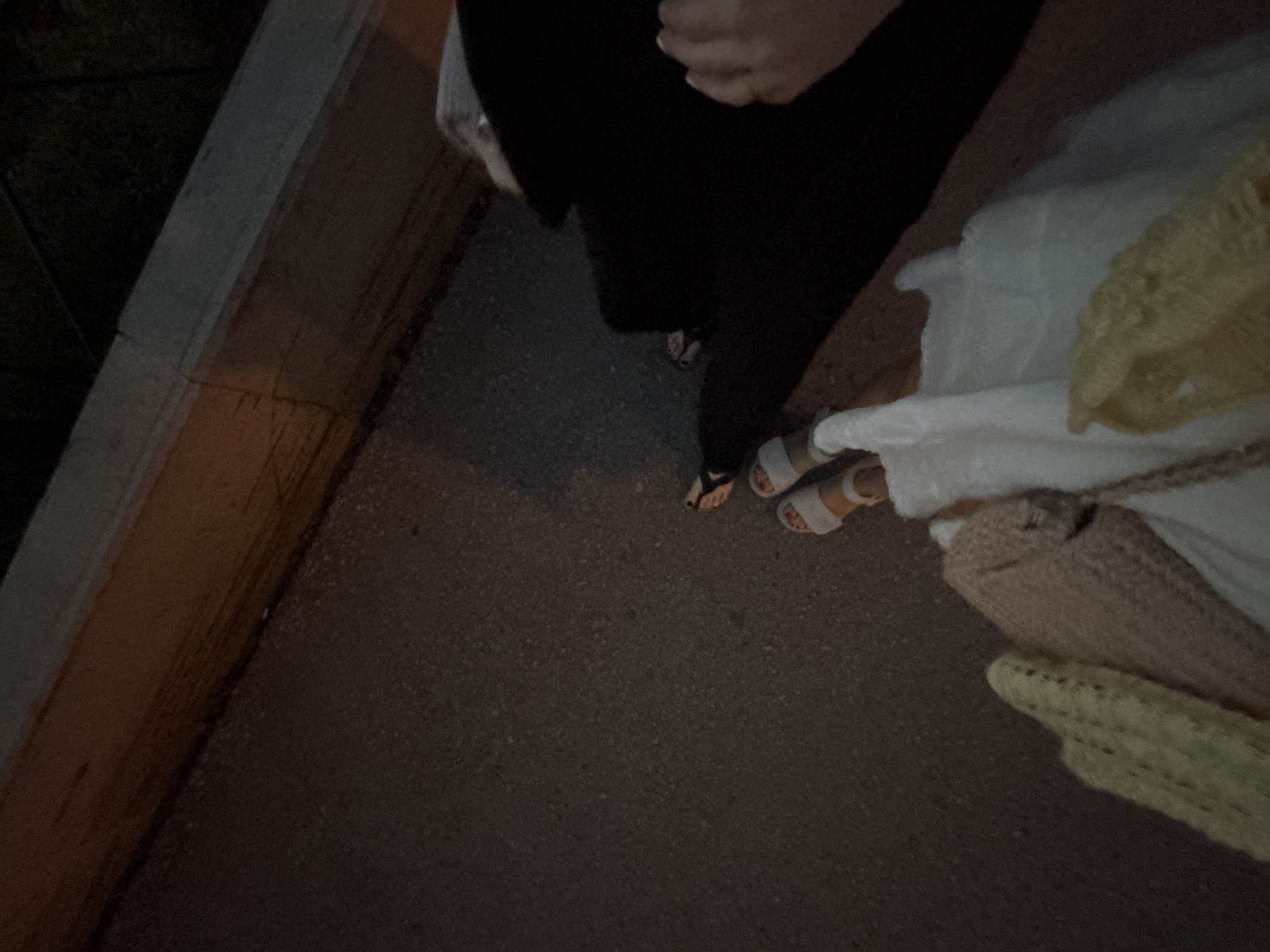 A high-angle, slightly blurred, low-light photograph depicts two individuals walking on a paved surface. The individual on the left wears dark-colored pants and open-toed sandals. The individual on the right wears white sandals and carries a white garment and a woven handbag. The setting appears to be a sidewalk adjacent to a concrete structure, possibly a bridge or elevated walkway. The location may be Perm, Russia, but this is not discernable from the visual evidence. No actions or interactions can be definitively determined beyond walking.