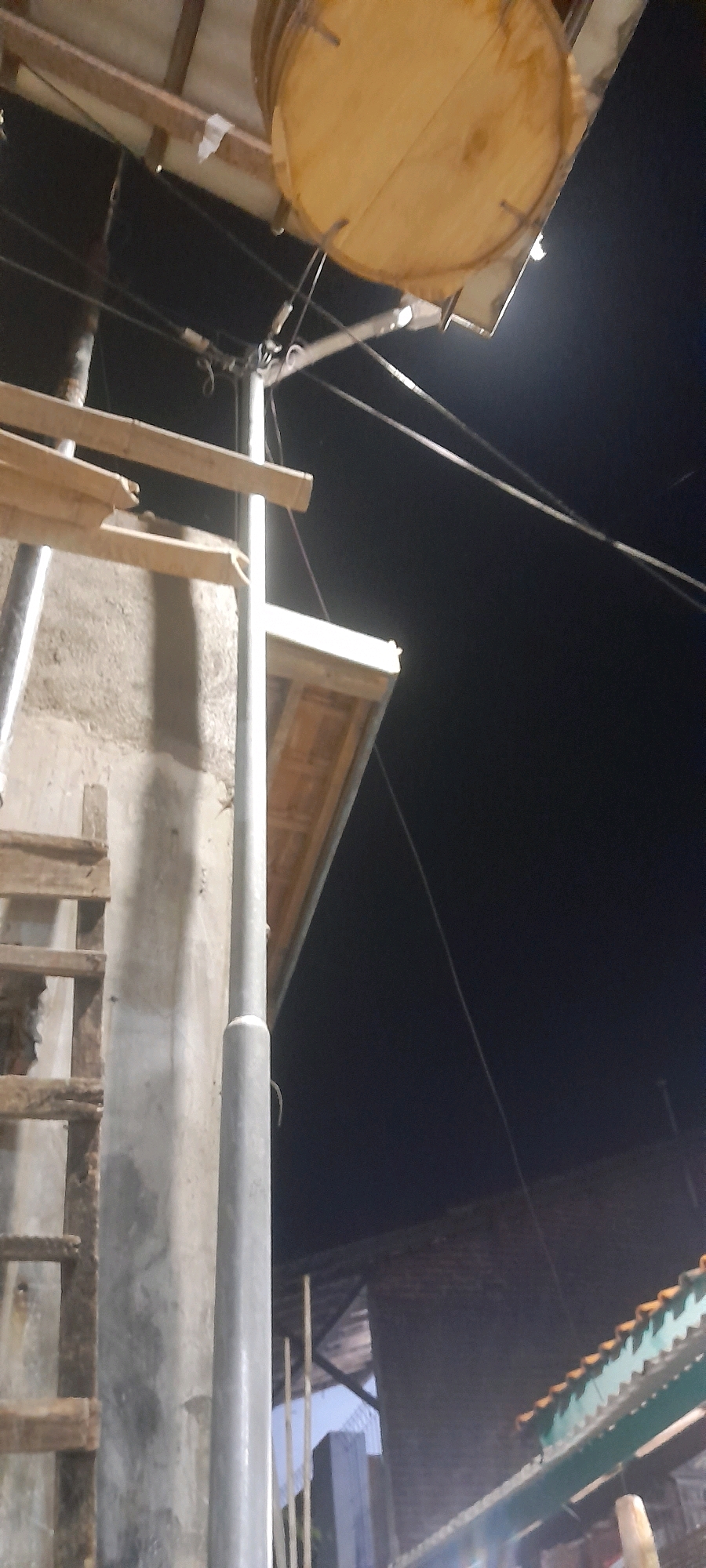 This image depicts an outdoor scene at night, likely in Tegal, Indonesia, with evidence of ongoing construction or renovation. A large, circular wooden object, possibly a drum or a large spool, is suspended from what appears to be a temporary roof structure made of wood. Below this, a gray utility pole stands prominently. To the left of the pole, a wooden ladder is attached to a rough concrete wall, suggesting access to a higher level or the roof. In the background, a dark sky contrasts with the illuminated structures, and the silhouette of a building with a corrugated roof is visible on the right. The overall atmosphere is dimly lit, indicative of nighttime conditions, with a focus on the construction elements.