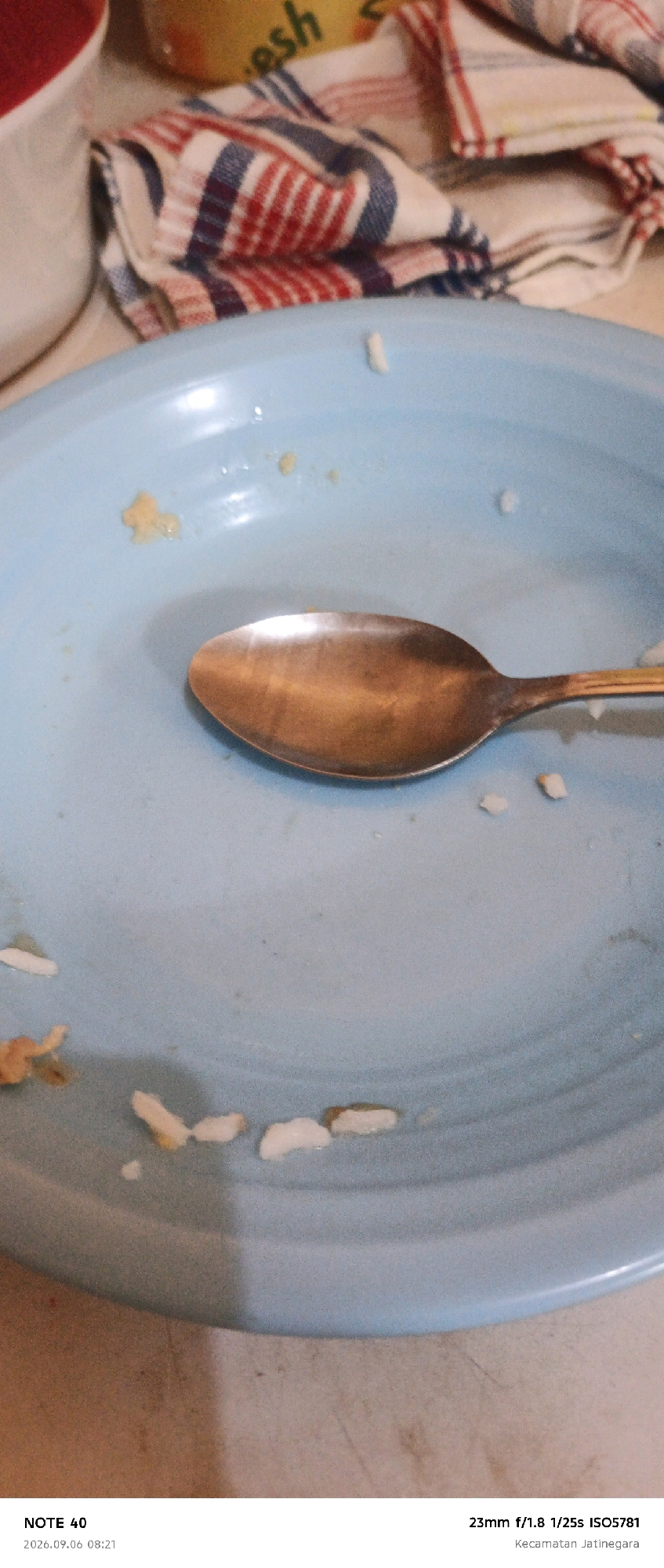 The image depicts a close-up, overhead view of a light blue plate with scattered rice grains and a single spoon. The plate is the primary subject, positioned in the lower half of the frame. A metallic spoon with a golden sheen rests on the plate, with its bowl facing upwards. Several white rice grains are dispersed across the plate's surface, with some appearing larger and clumped, while others are small and individual. In the upper portion of the image, a folded red, white, and blue striped cloth is visible, along with a blurry yellow object. The background consists of a light brown surface. Text at the bottom of the image indicates "NOTE 40", a date and time "2026.09.06 08:21", and camera settings "23mm f/1.8 1/25s ISO5781", along with "Kecamatan Jatinegara", which is a subdistrict in East Jakarta, Indonesia.
