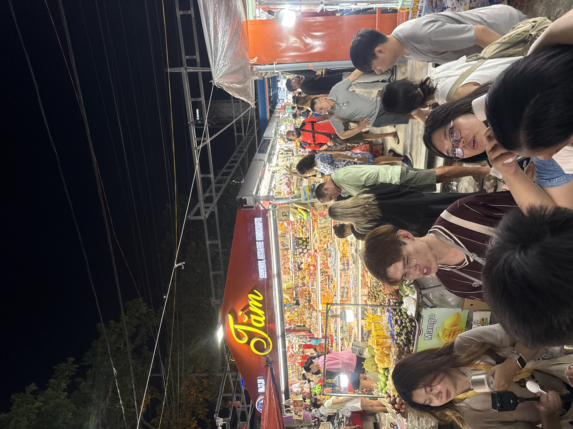 The image shows a bustling night market scene in an unknown city. The foreground is filled with people, many of whom appear to be Asian, looking at various items for sale. There are some who are looking at mangoes and durians. Overhead lights illuminate the market, and a sign with the name "Jam" is visible in large, illuminated letters. The sign also mentions "SOUVENIRS, ARTS&FINE HANDICRAFTS". Various goods are displayed on shelves and tables. In the background, more people are visible, moving through the market. The night sky is dark, but power lines are visible running along the top left of the frame.