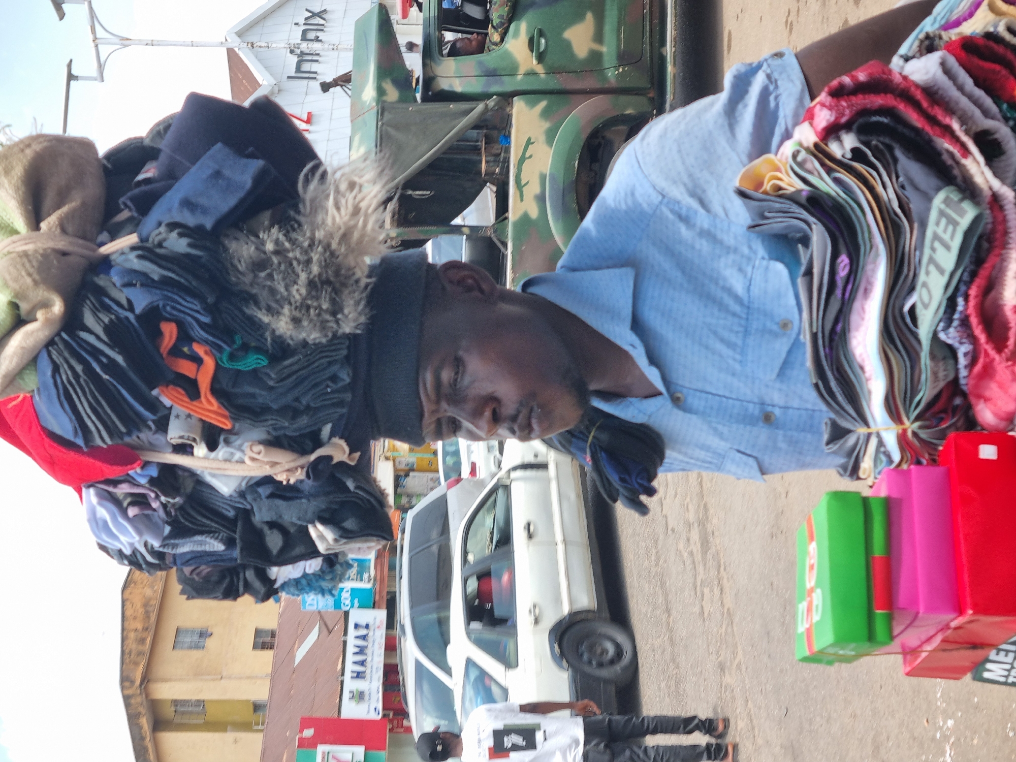 A man stands outdoors, carrying a large bundle of clothing on his head and more in his arms. He wears a blue button-down shirt and a black beanie, while the clothing appears to be a mix of colors and textures. Behind him, an olive-green military vehicle is partially visible. Further in the background, a white van and other vehicles are parked along a street. Buildings are visible, one with a sign that reads "HAMAZ" on the street. The scene is set on the city of Jos, Nigeria.